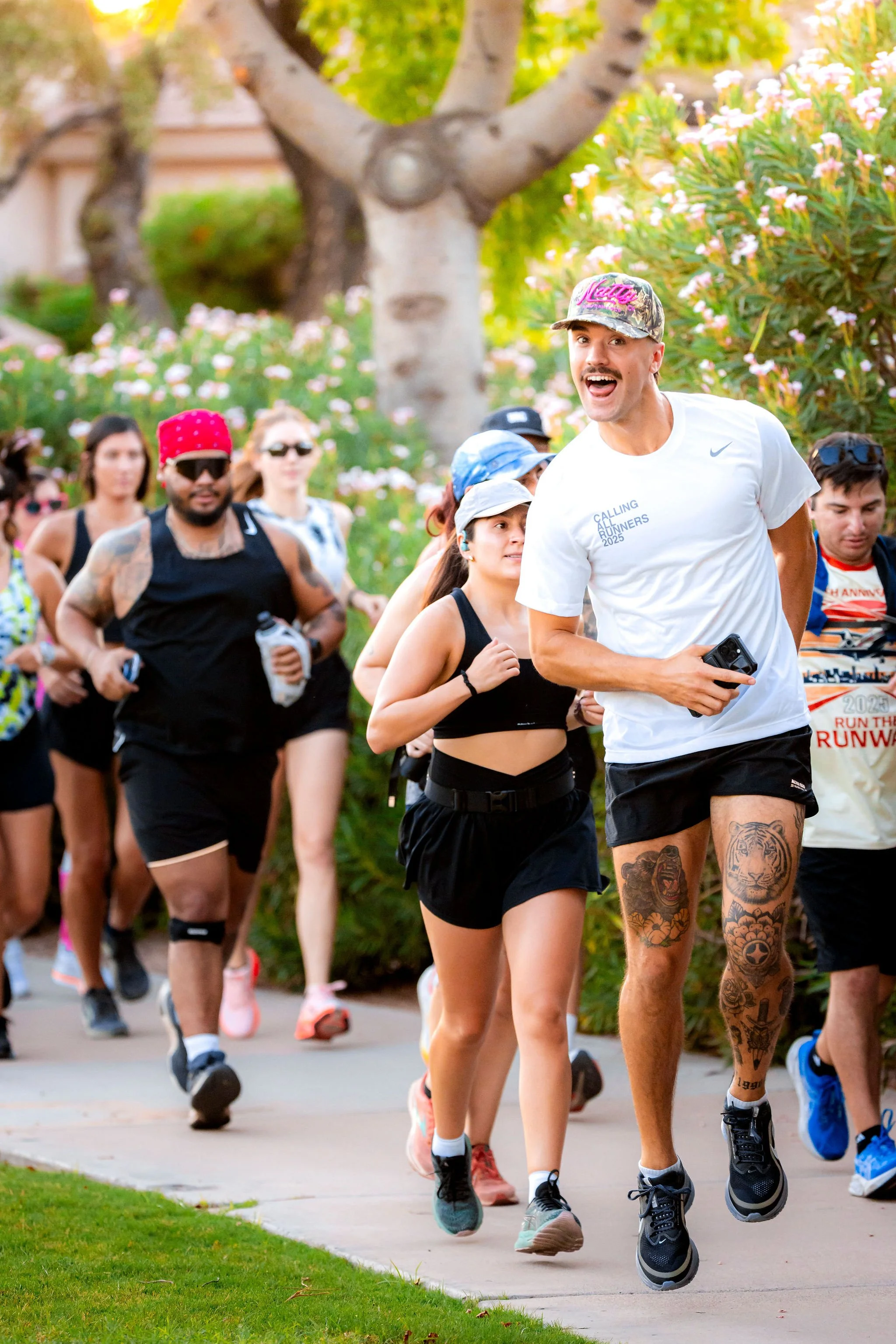 A group of people participating in a run or marathon outdoors in a lush park setting, with trees and flowering bushes in the background.