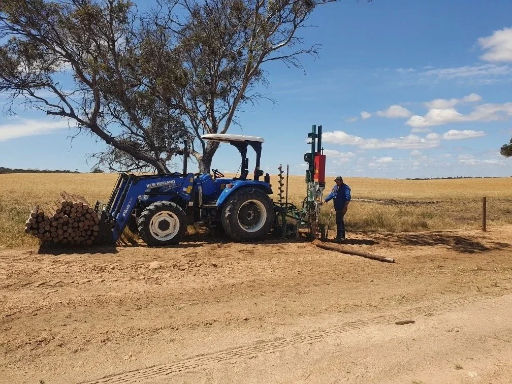 A tractor on a dirt road pulling logs with a worker in blue operating a drilling machine next to a field of yellow crops and a tree under a partly cloudy sky.