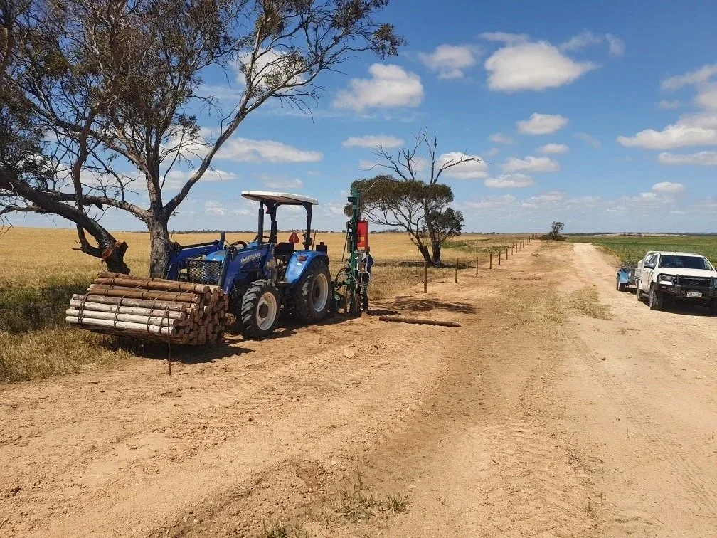 A tractor hauling logs parked on a dirt path next to a tree and open fields under a cloudy sky.