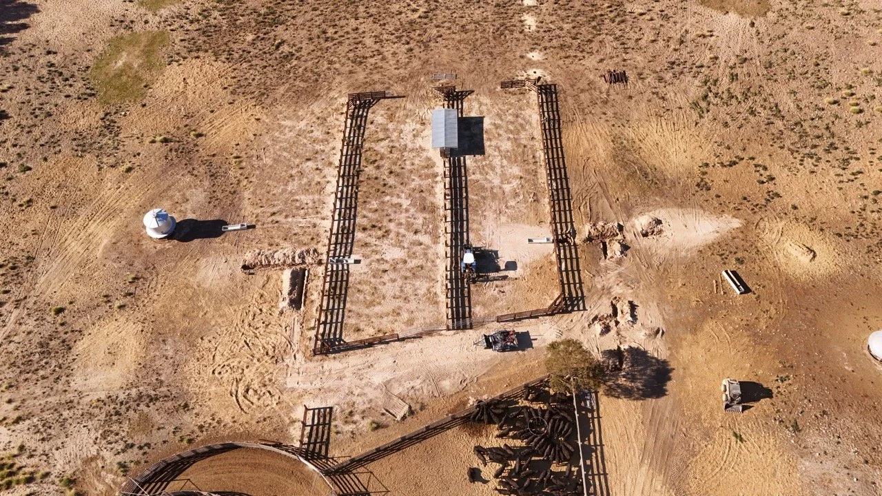Aerial view of a desert area with a fenced enclosure, a small building, and various equipment and structures, including water tanks and a solar panel.