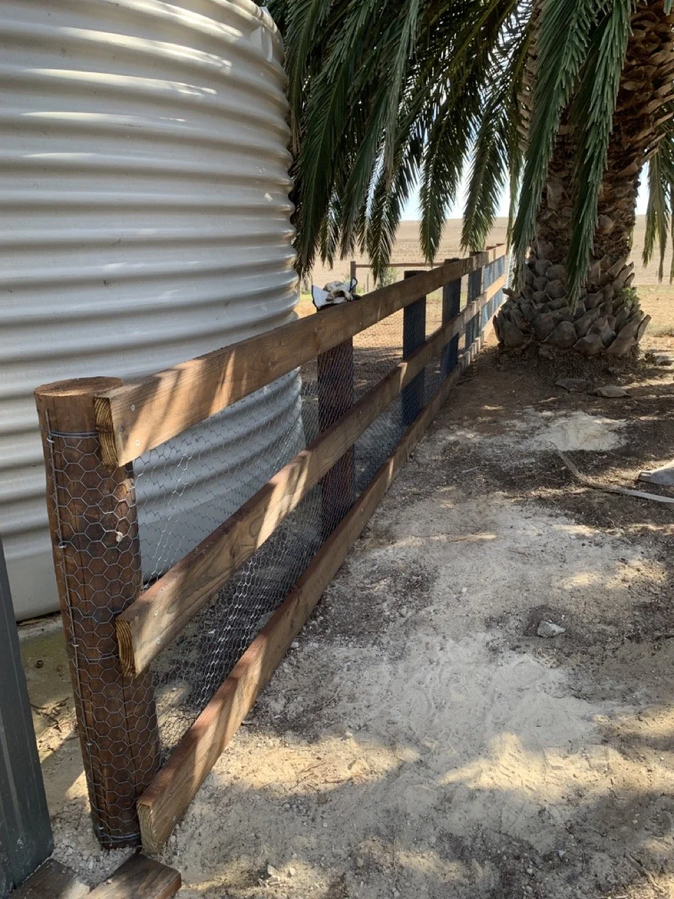 A wooden and wire fence running beside a large palm tree and a metal water tank in an outdoor setting.