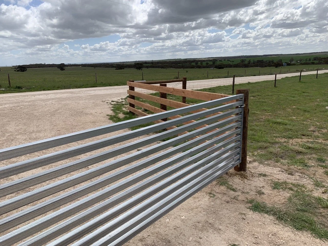 A metal gate in the foreground with a wooden fence behind it, opening to a dirt road and open green fields under a partly cloudy sky.