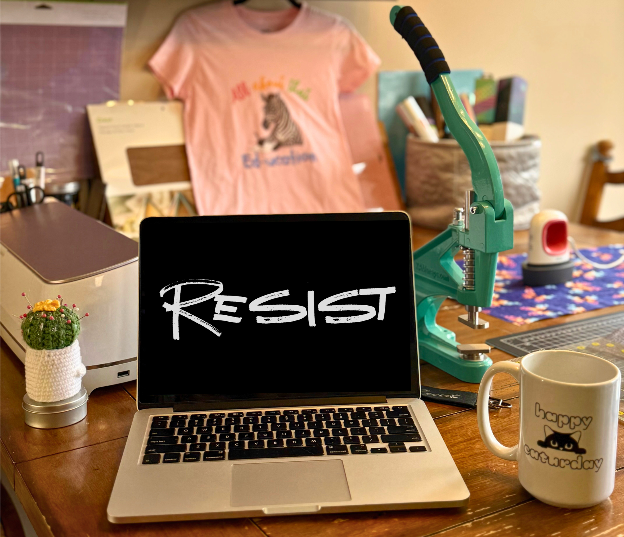 Desk scene with a laptop displaying 'RESIST' on the screen, a coffee mug with a cat and 'happy caturday' text, a small plant in a knitted cover, a professional teal snap press, and various office supplies and decorations in the background.