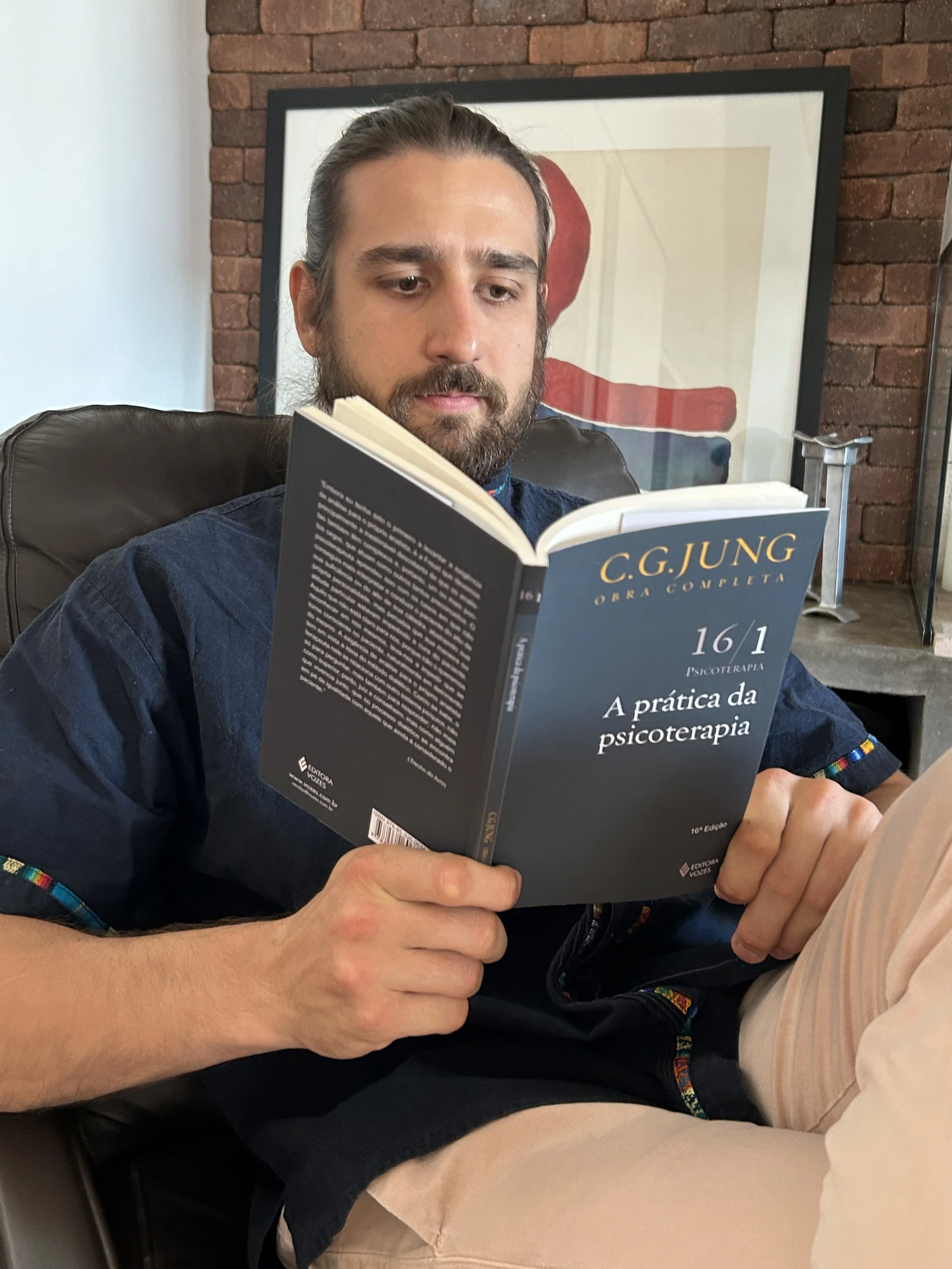Homem com cabelo preso e barba, lendo um livro azul intitulado 'C.G. Jung obra completa 16/1 A prática da psicoterapia', sentado em uma cadeira de couro, com fundo de parede de tijolos e uma obra de arte atrás dele.
