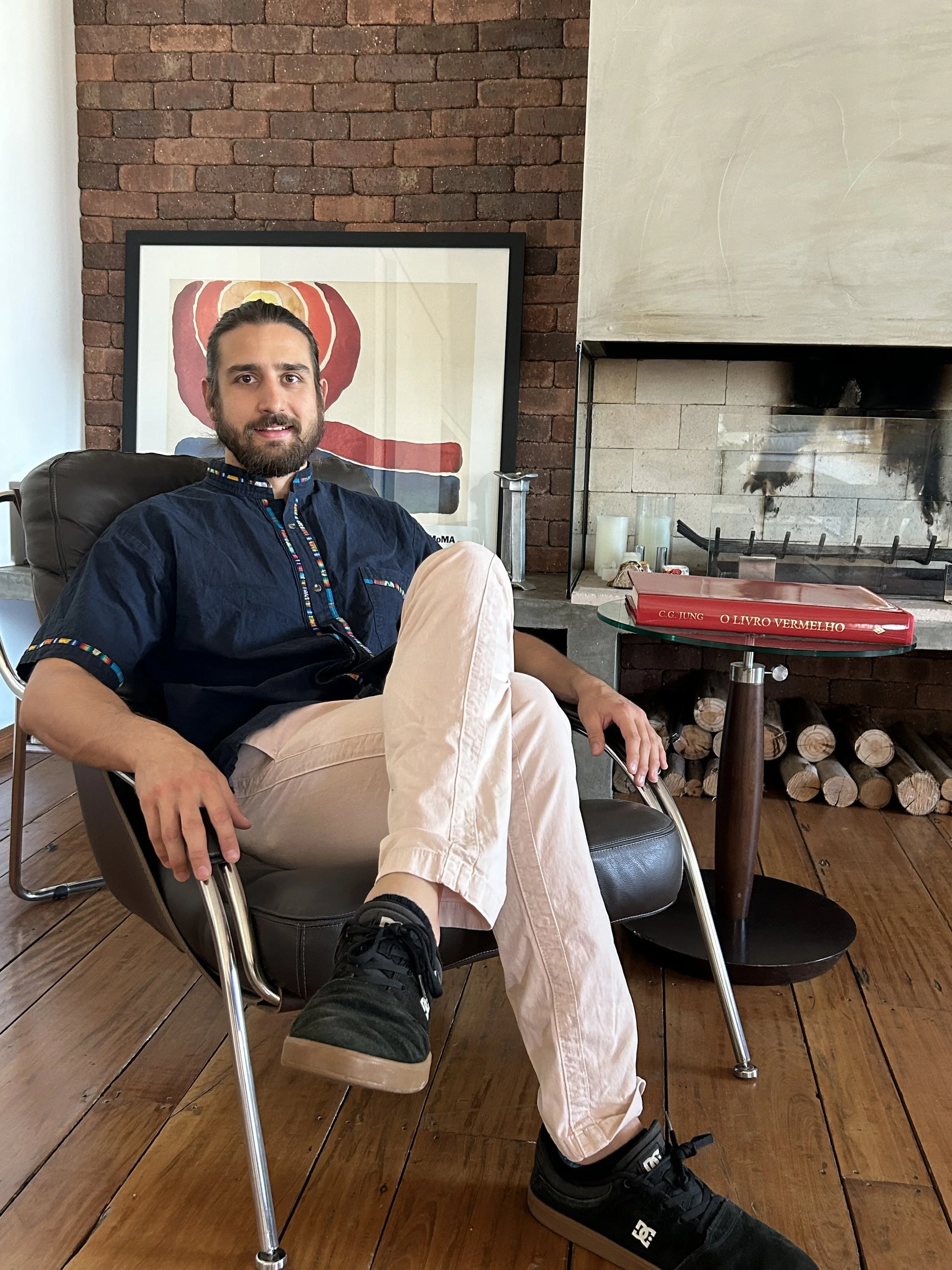 Homem sentado em uma cadeira de couro preto, com pernas cruzadas, usando camiseta azul, calça bege e tênis preto, em um ambiente com parede de tijolos ao fundo, uma obra de arte, uma lareira e uma mesa com uma pasta vermelha sobre ela.