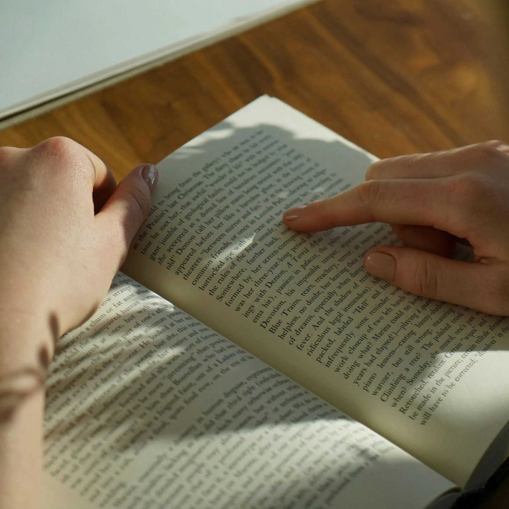Pessoa segurando um livro aberto, com o foco na página de textos, em uma superfície de madeira.