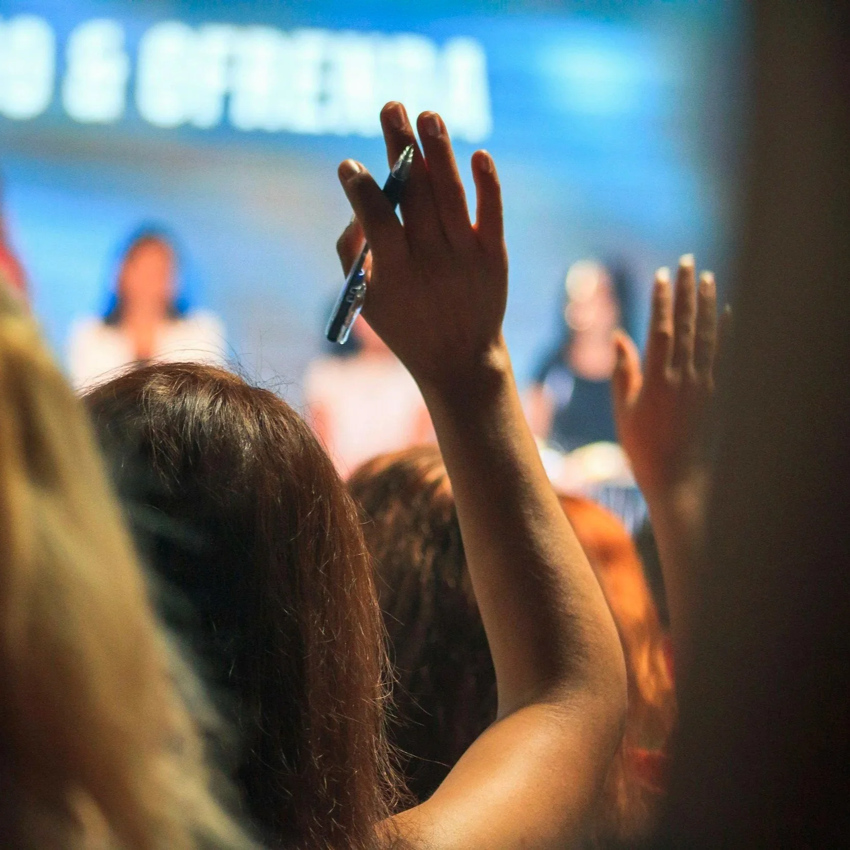 Pessoas levantando as mãos em um evento, com uma pessoa segurando uma caneta.