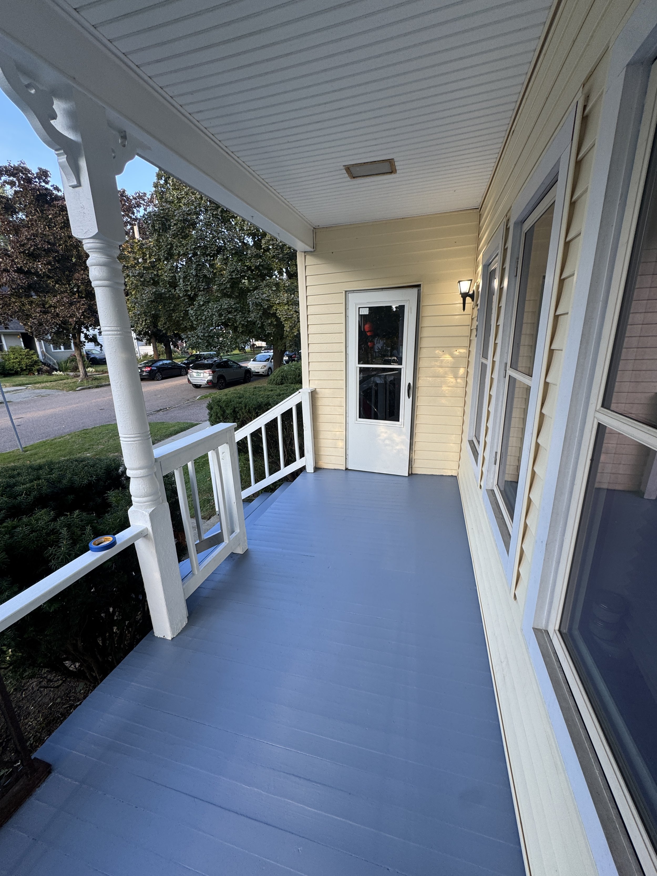 A covered porch with light yellow siding, a white door with a window, a wall-mounted light fixture, and white railing. The porch has a blue wooden floor and overlooks a street with parked cars and trees.