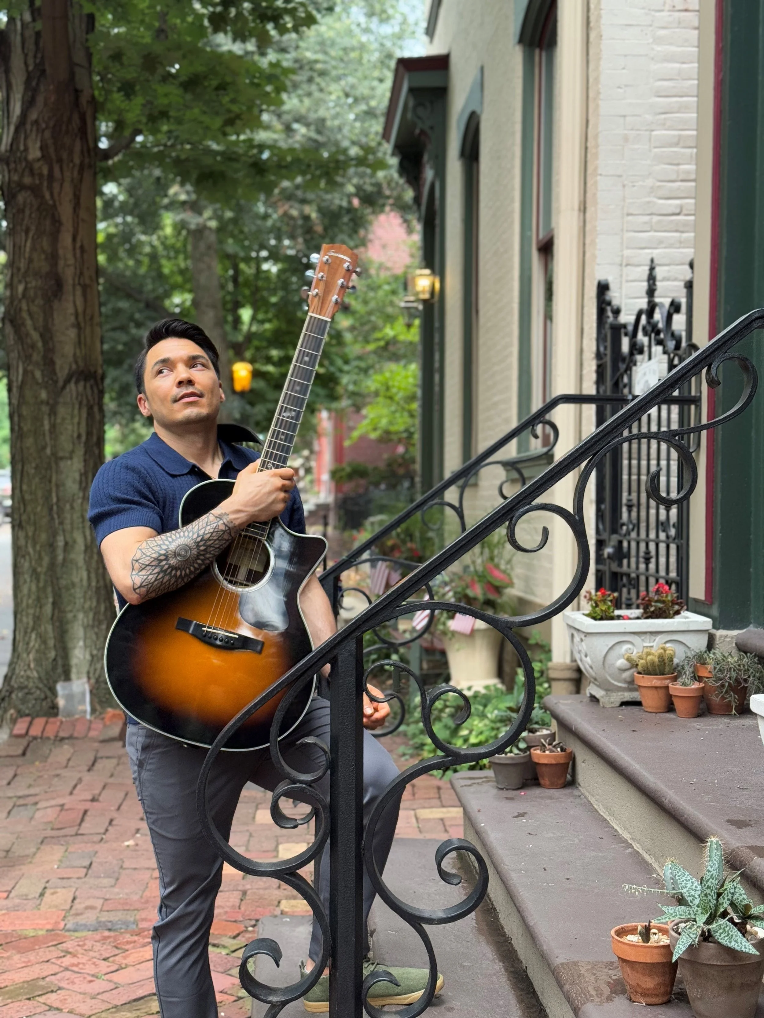 Man with tattoos playing an acoustic guitar on a brick sidewalk next to stairs with potted plants, near a building with ornate iron railings and surrounded by green trees.