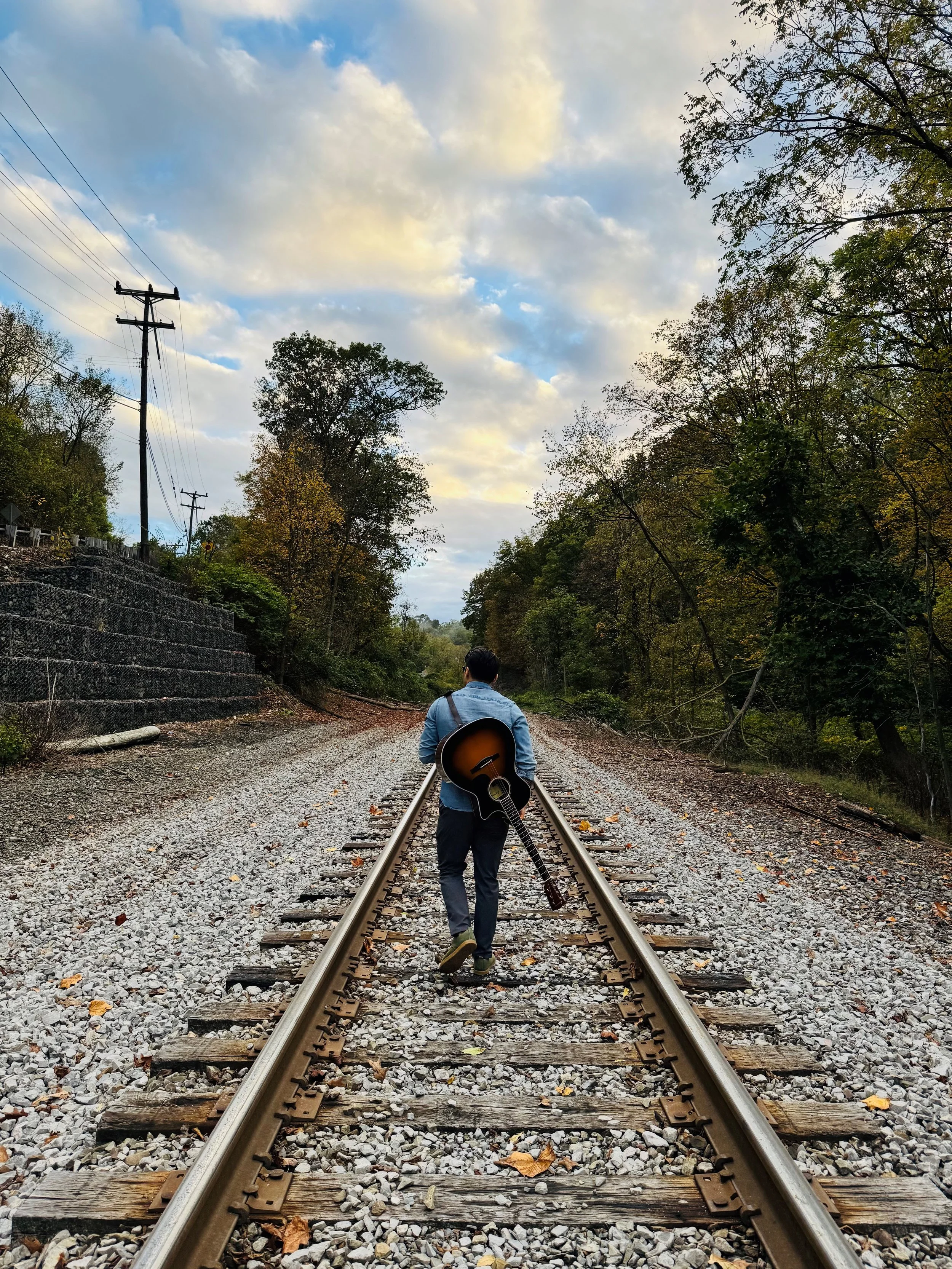A person walking along railroad tracks carrying a guitar, with trees and a cloudy sky in the background.