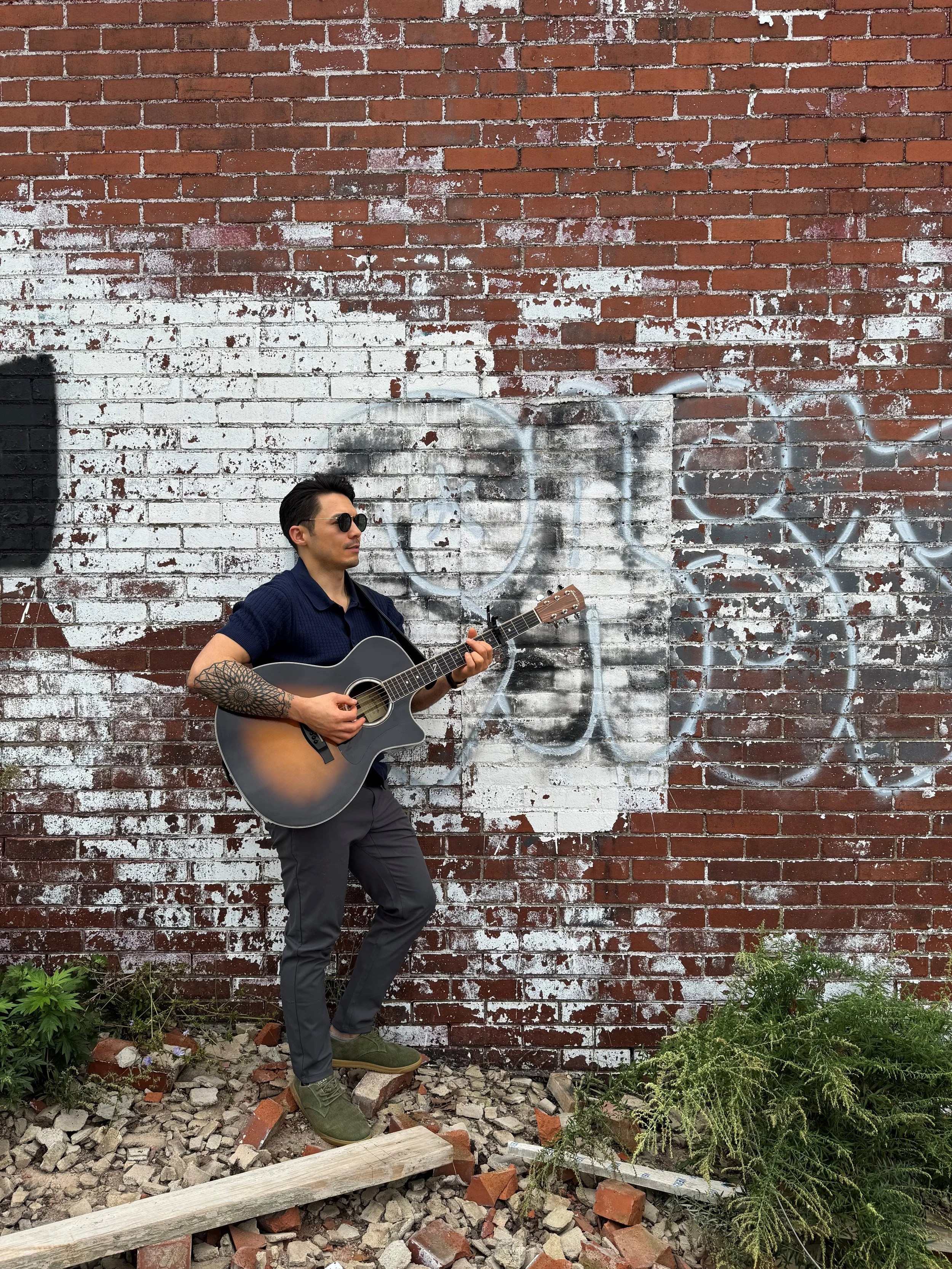 A man with sunglasses playing an acoustic guitar against a brick wall with graffiti. The ground is covered with rubble and there is some greenery.
