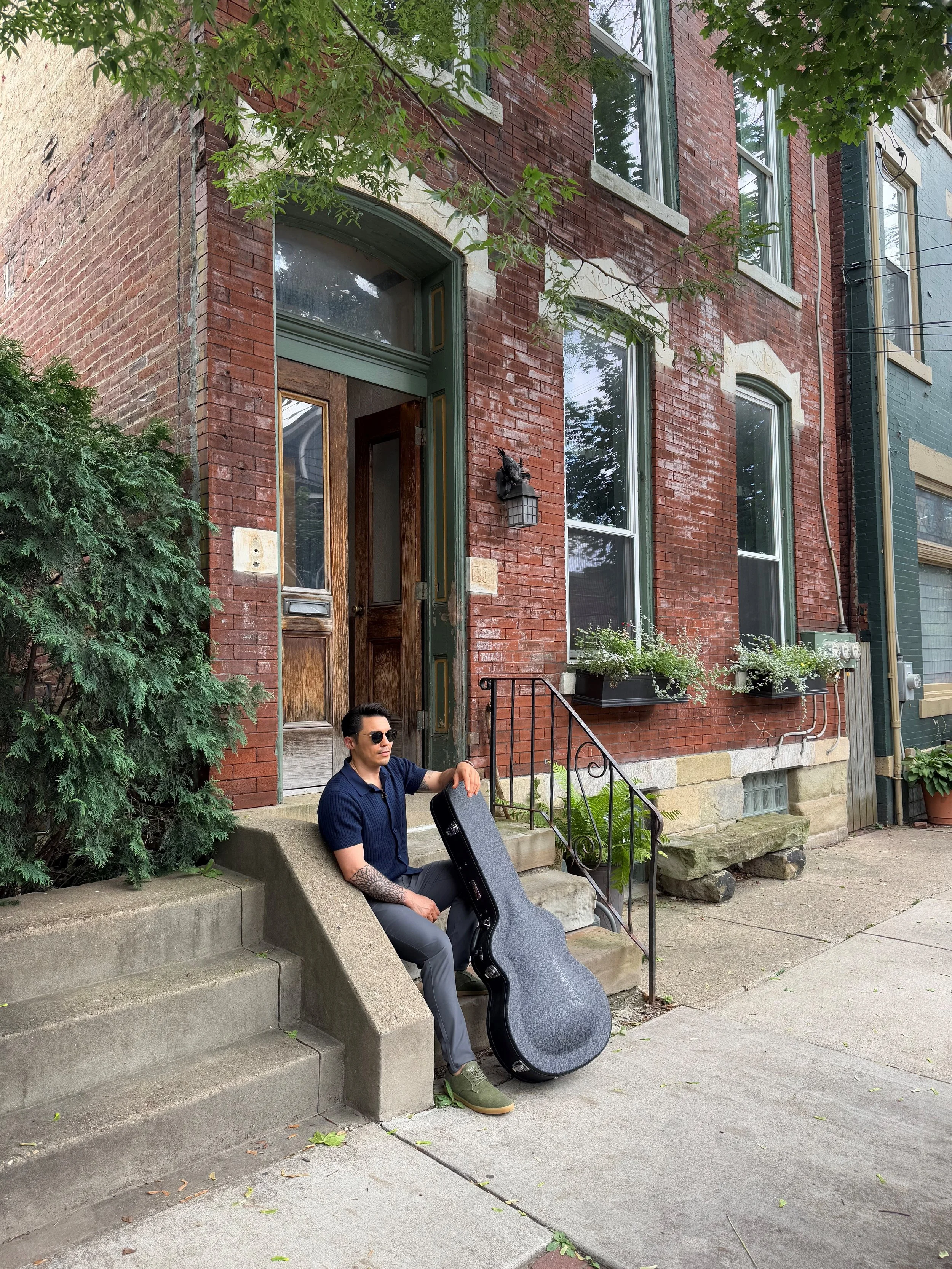 A man wearing sunglasses, a dark blue shirt, and gray pants sitting on the concrete stairs outside a red brick building, holding a guitar case. There are green plants, a window with window boxes, and a tree in the background.