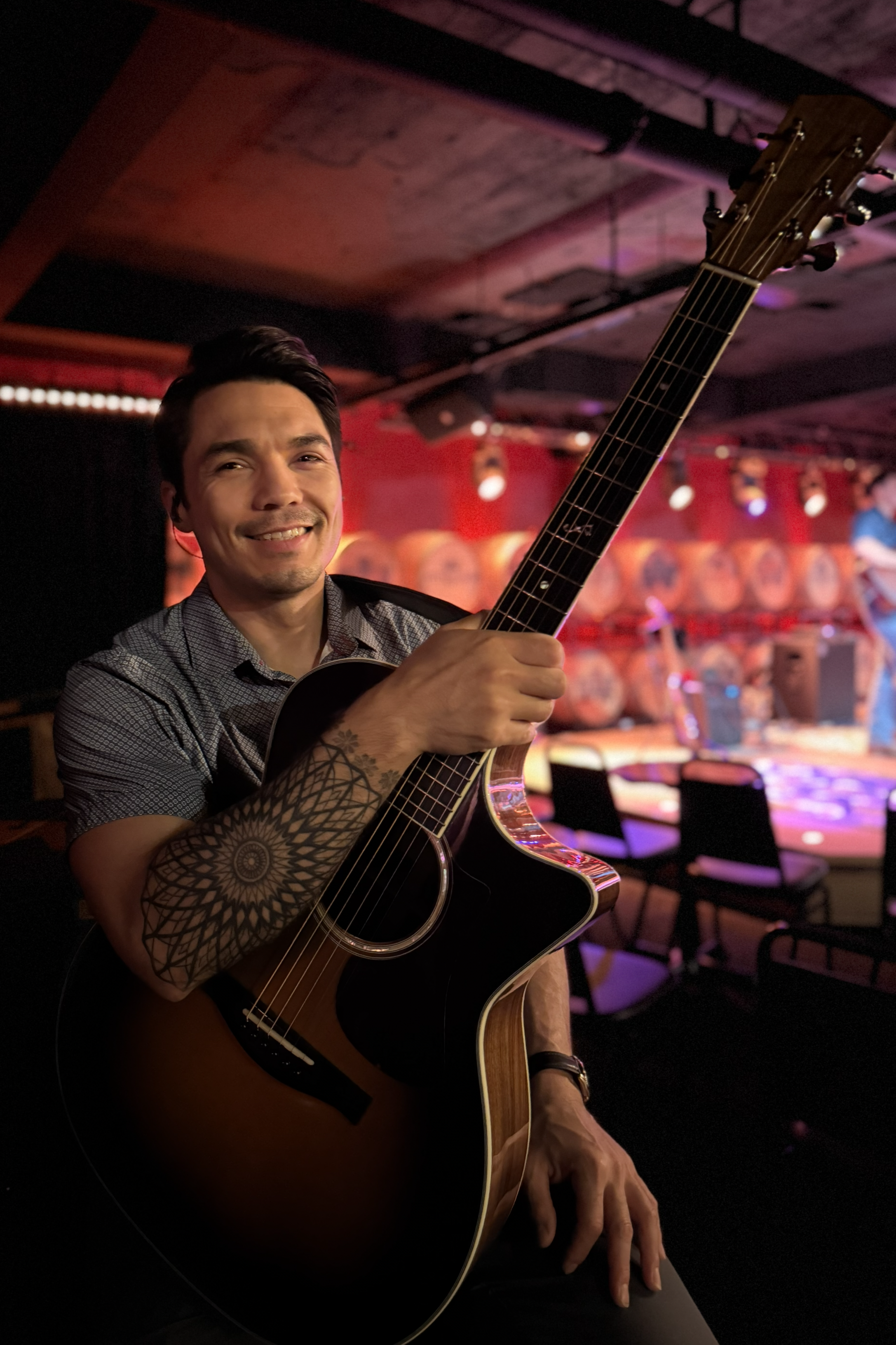 Kaye Baron smiling while holding an acoustic guitar, sitting in a dimly lit venue with a stage in the background featuring a red backdrop and barrels.