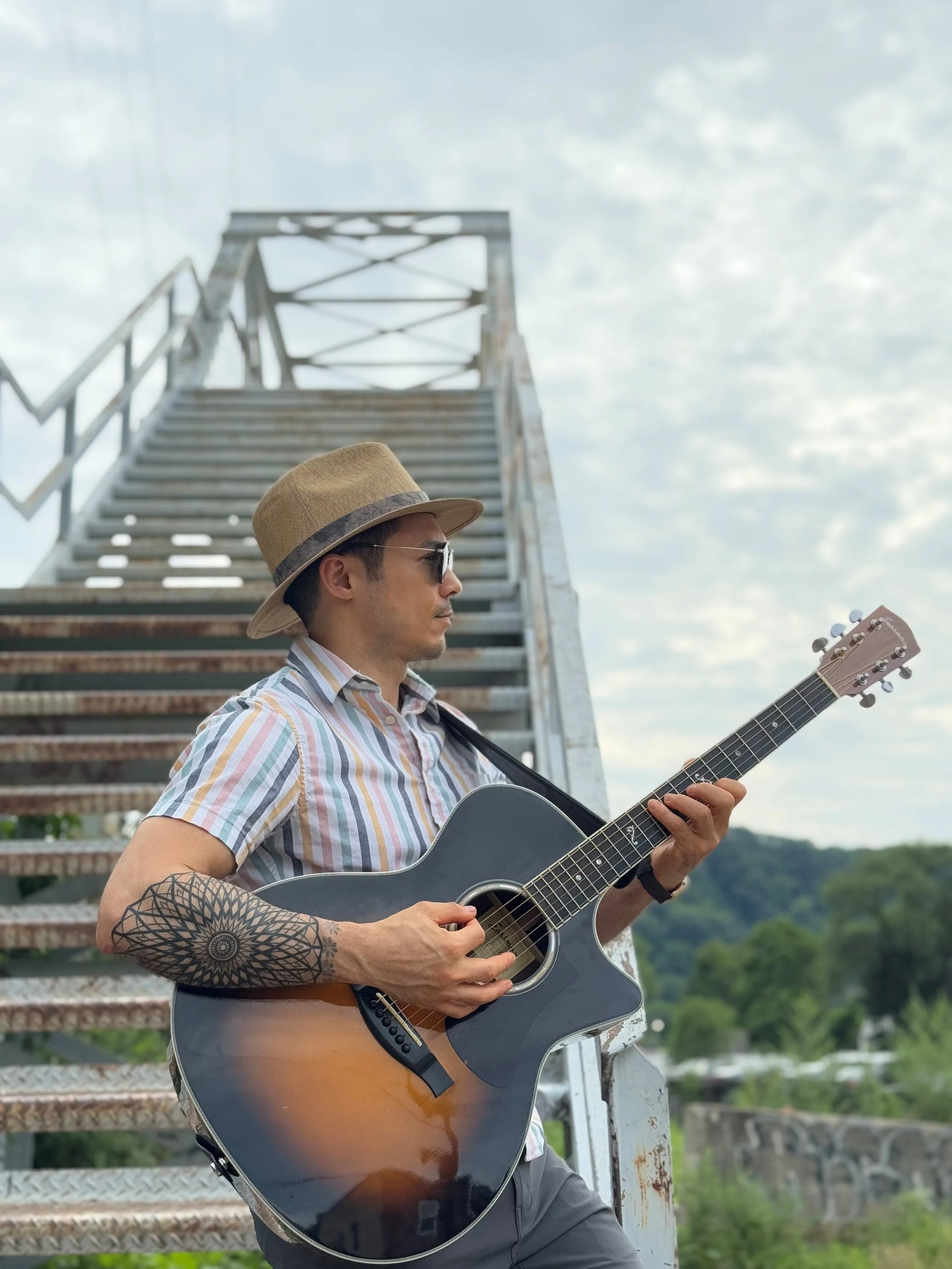 A man wearing sunglasses, a striped shirt, a hat, and a tattoo on his arm is playing an acoustic guitar outdoors near a metal staircase with a cloudy sky and green landscape in the background.