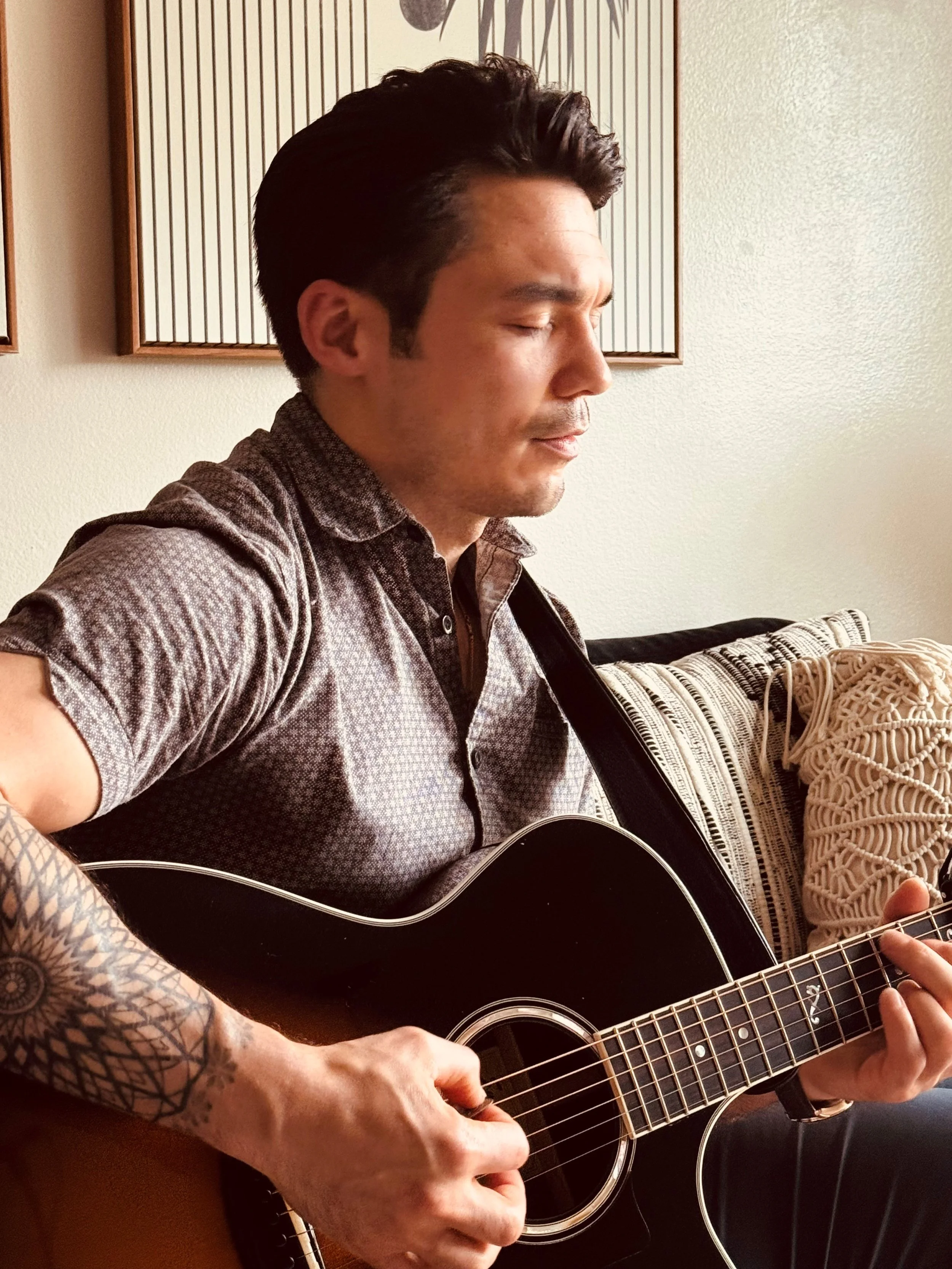 A man with dark hair and a tattoo on his left arm playing a black acoustic guitar with his eyes closed in a cozy indoor setting.