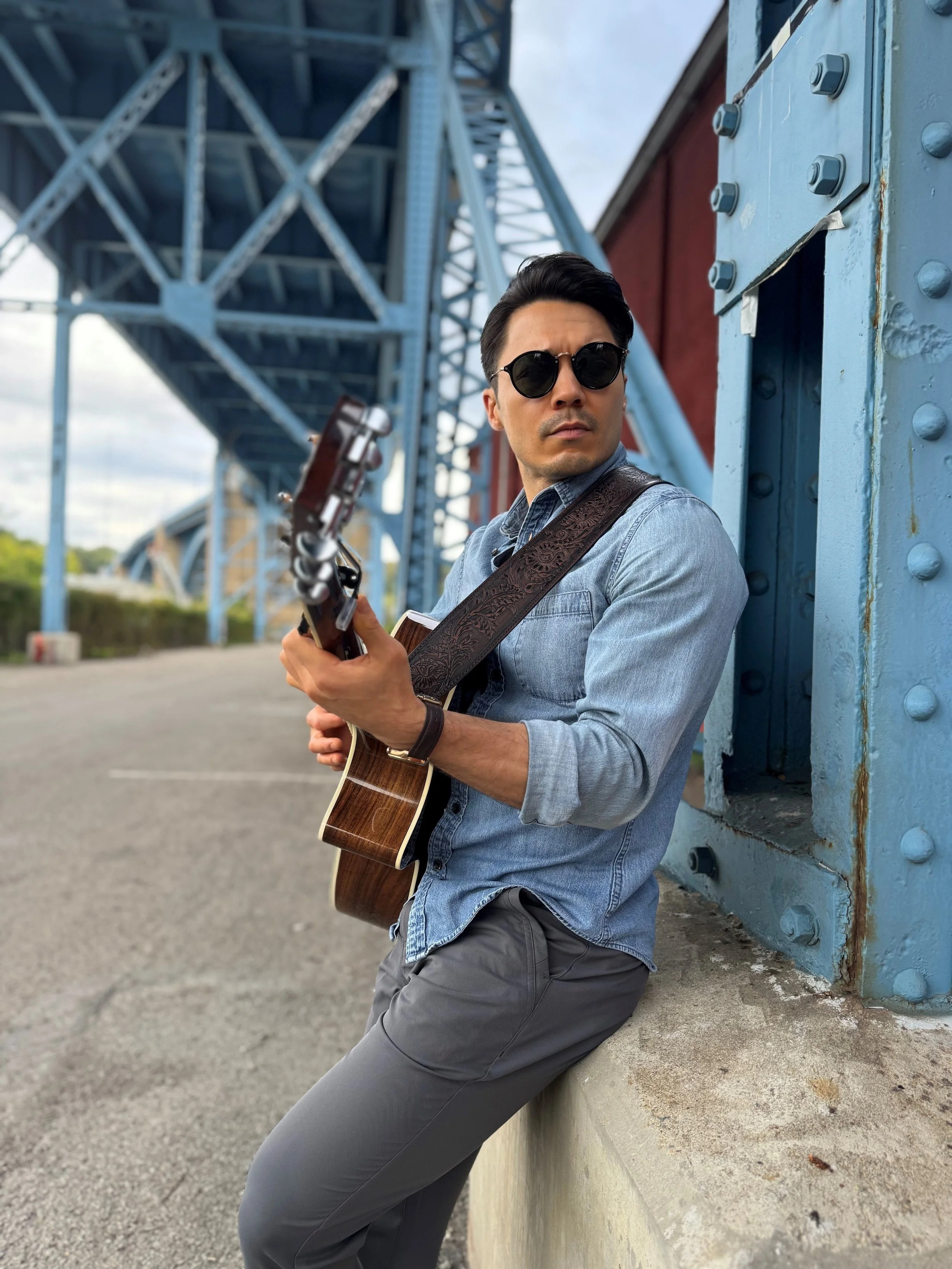 A man wearing sunglasses, a denim shirt, and gray pants, playing an acoustic guitar outdoors near an industrial-looking blue metal structure, possibly a bridge or pier.