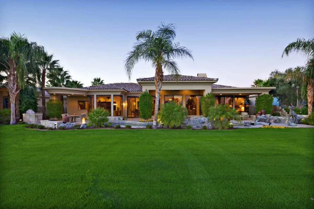 A large house with a landscaped backyard that features palm trees, shrubs, rocks, and a seating area, with warm interior lighting visible through large windows.