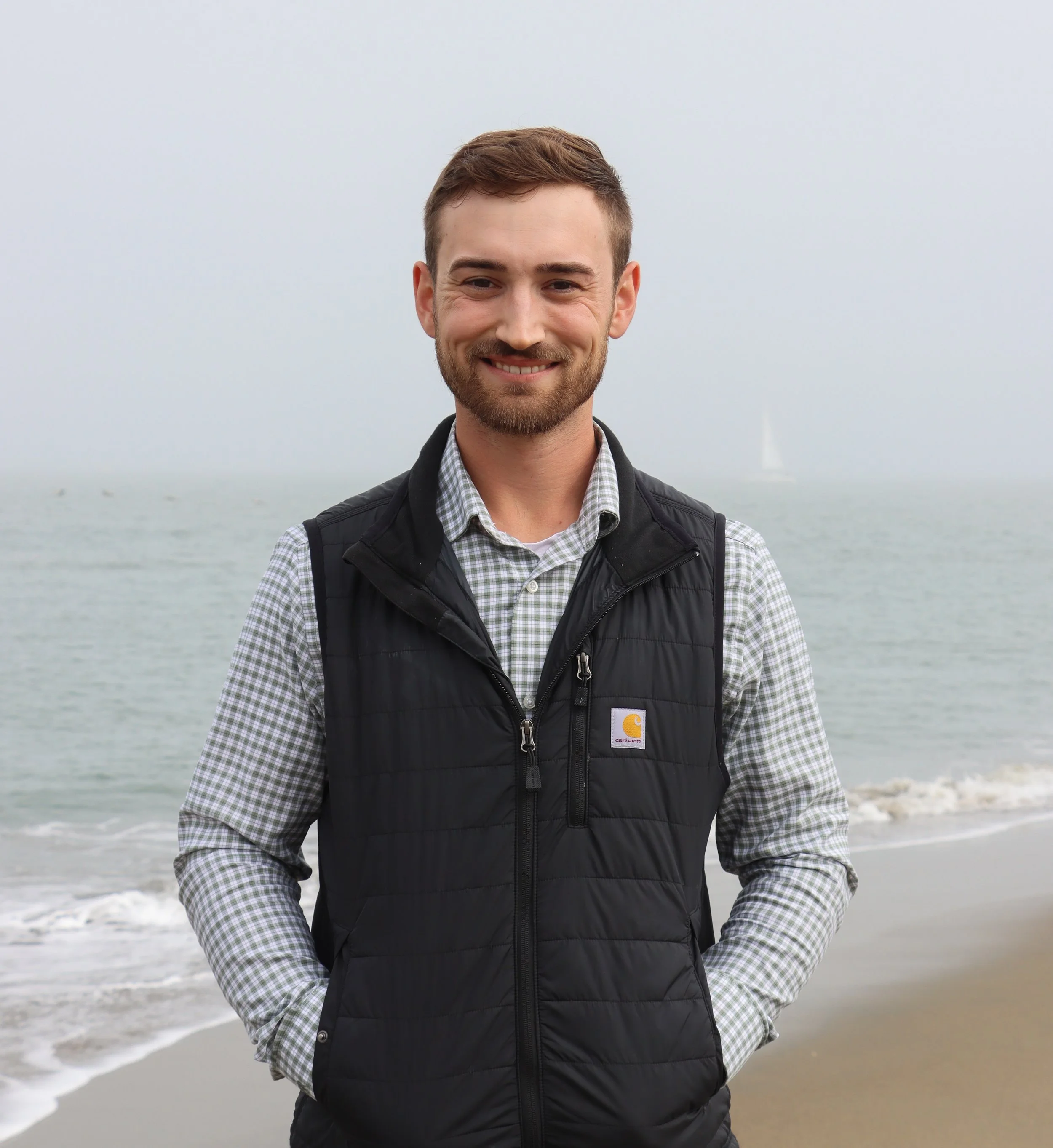 A man with a beard and short brown hair smiling at the camera on a foggy beach, wearing a checkered shirt and a black vest.