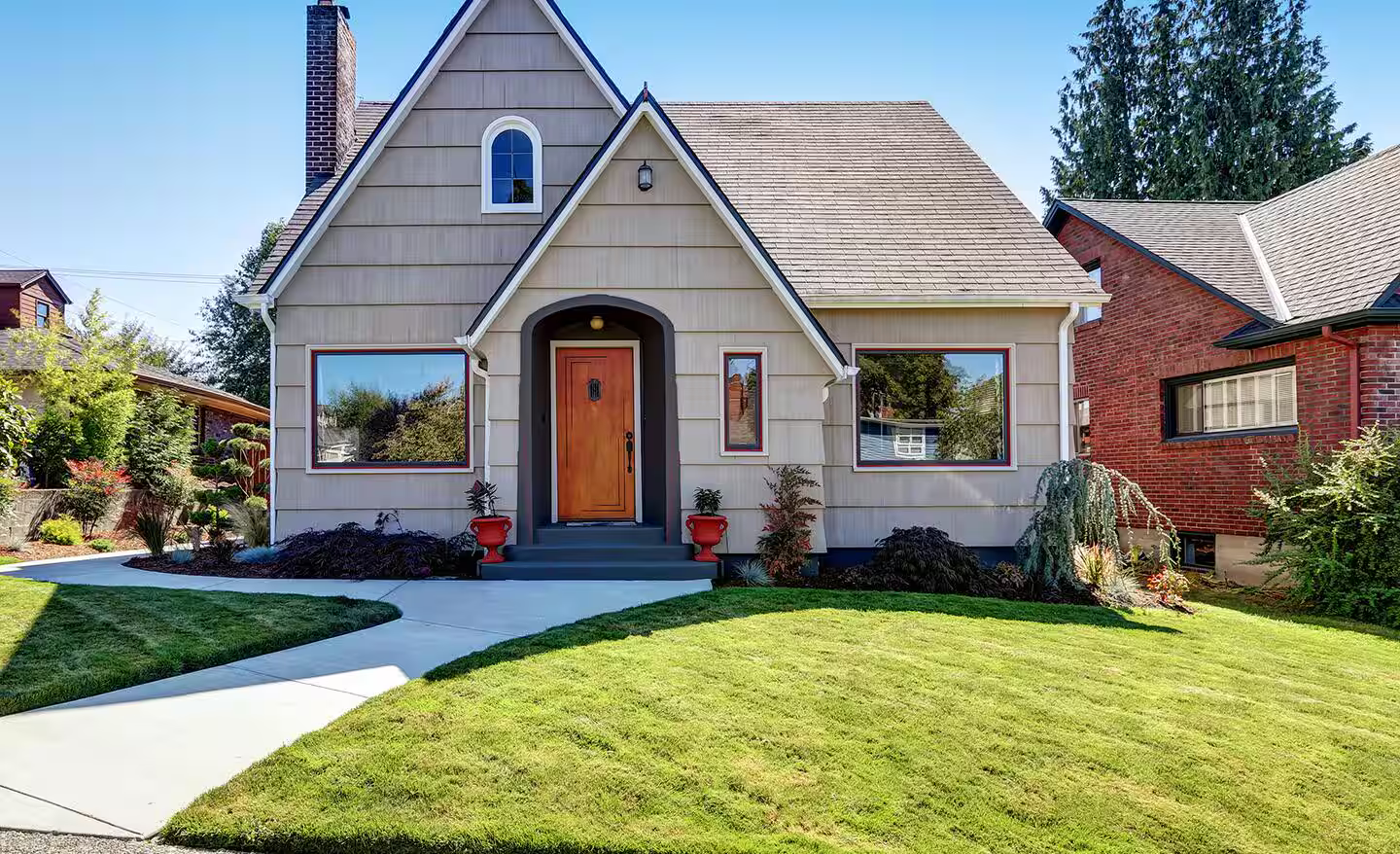 Front view of a house with a gable roof, beige siding, a wooden front door, large windows, and a well-maintained green lawn.