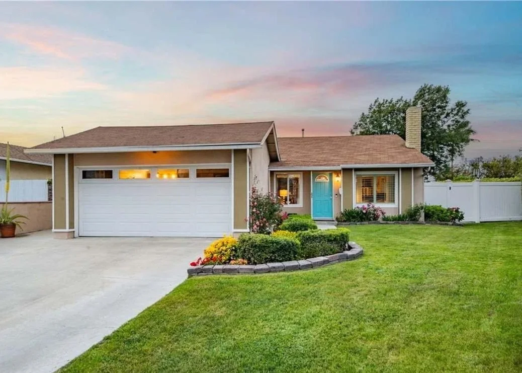 Front view of a single-story house with a blue front door, a white garage door, a manicured lawn, and a flower bed at sunset.