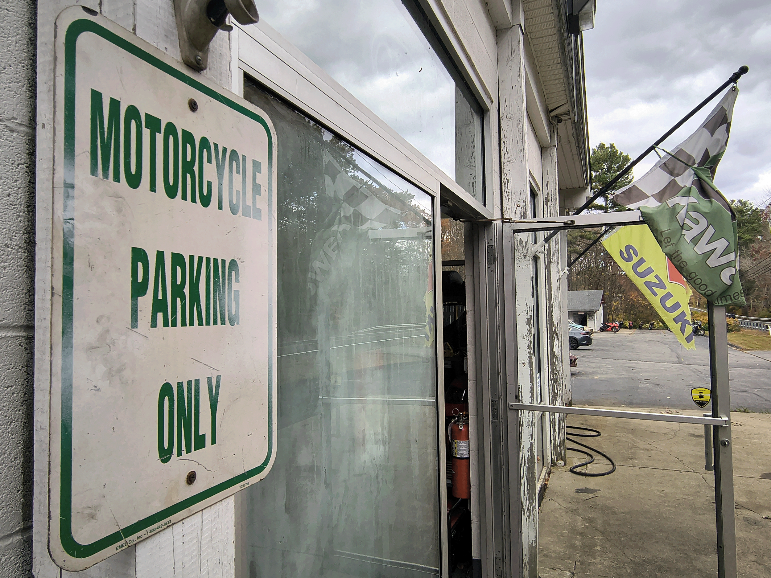 A sign on a wall reads 'Motorcycle Parking Only'. There are flags advertising various brands, including Suzuki, hanging outside a building with a glass window and an open door, in a parking lot with some cars and motorcycles visible in the background.