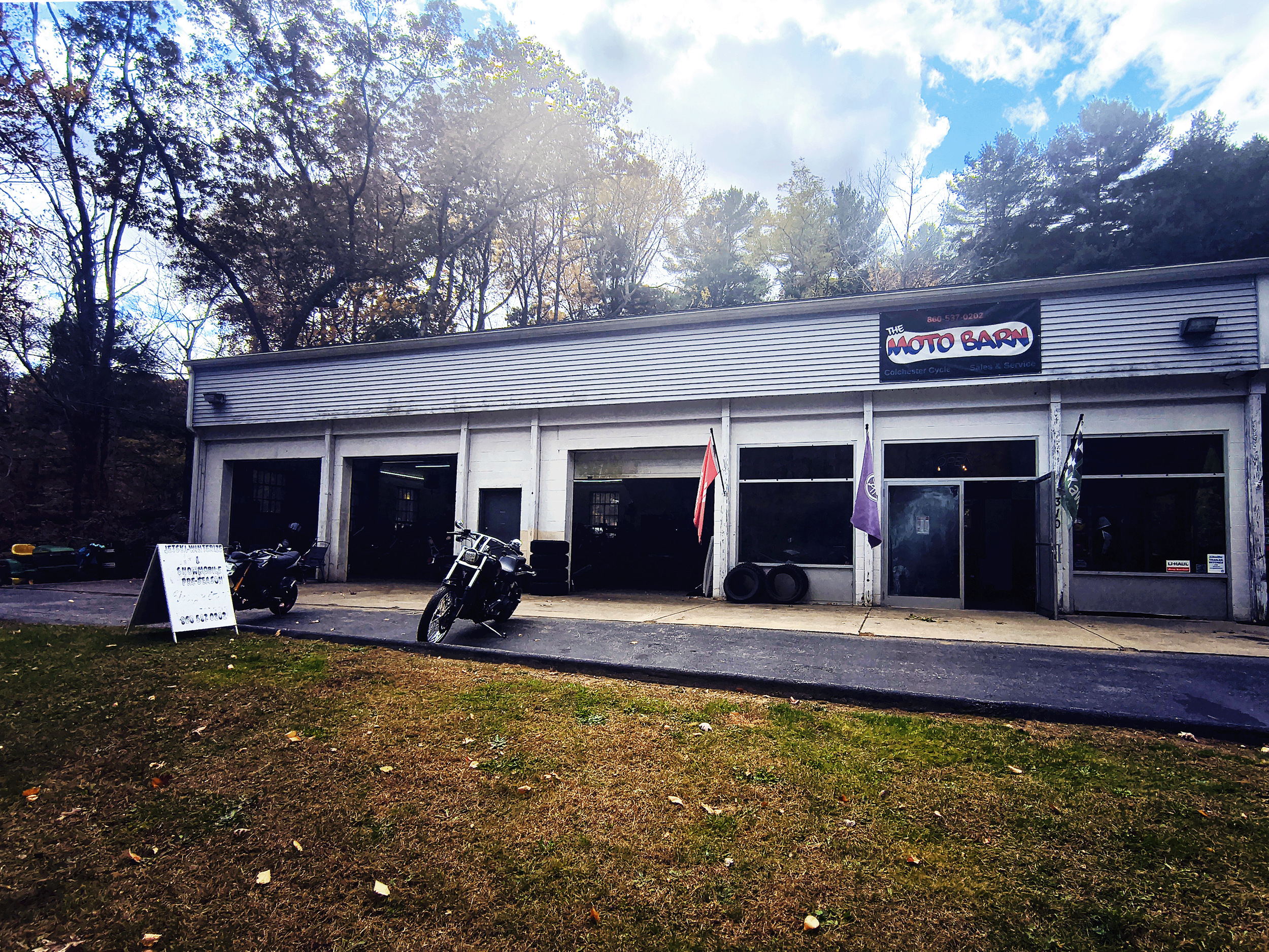 The Moto Barn, a motorcycle dealership with an open garage, motorcycles parked outside, and flags displayed in front, situated in a wooded area.