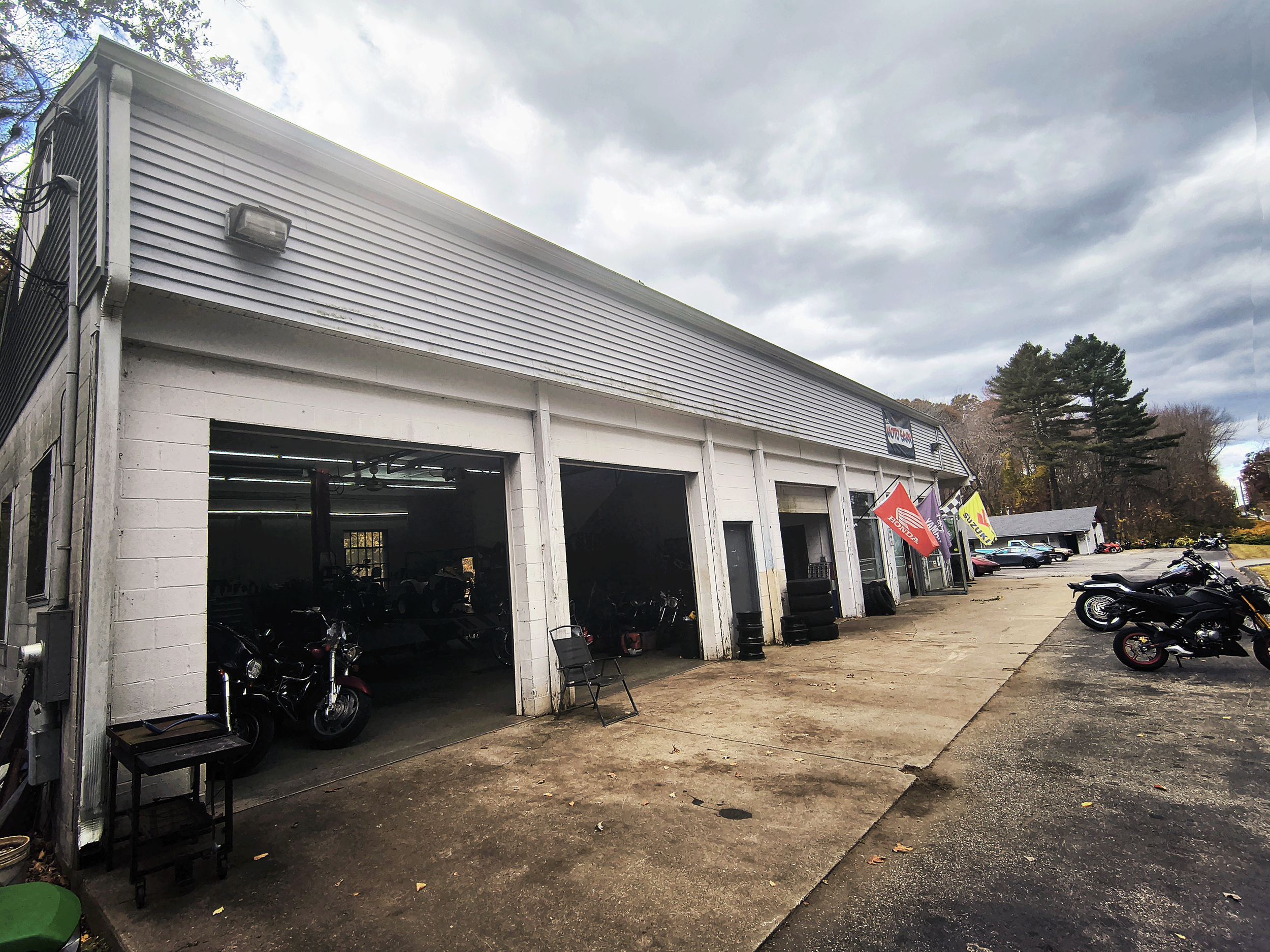 A motorcycle shop with open garage bays displaying motorcycles inside and outside, flags on the front, and a parking lot with motorcycles and cars under a cloudy sky.