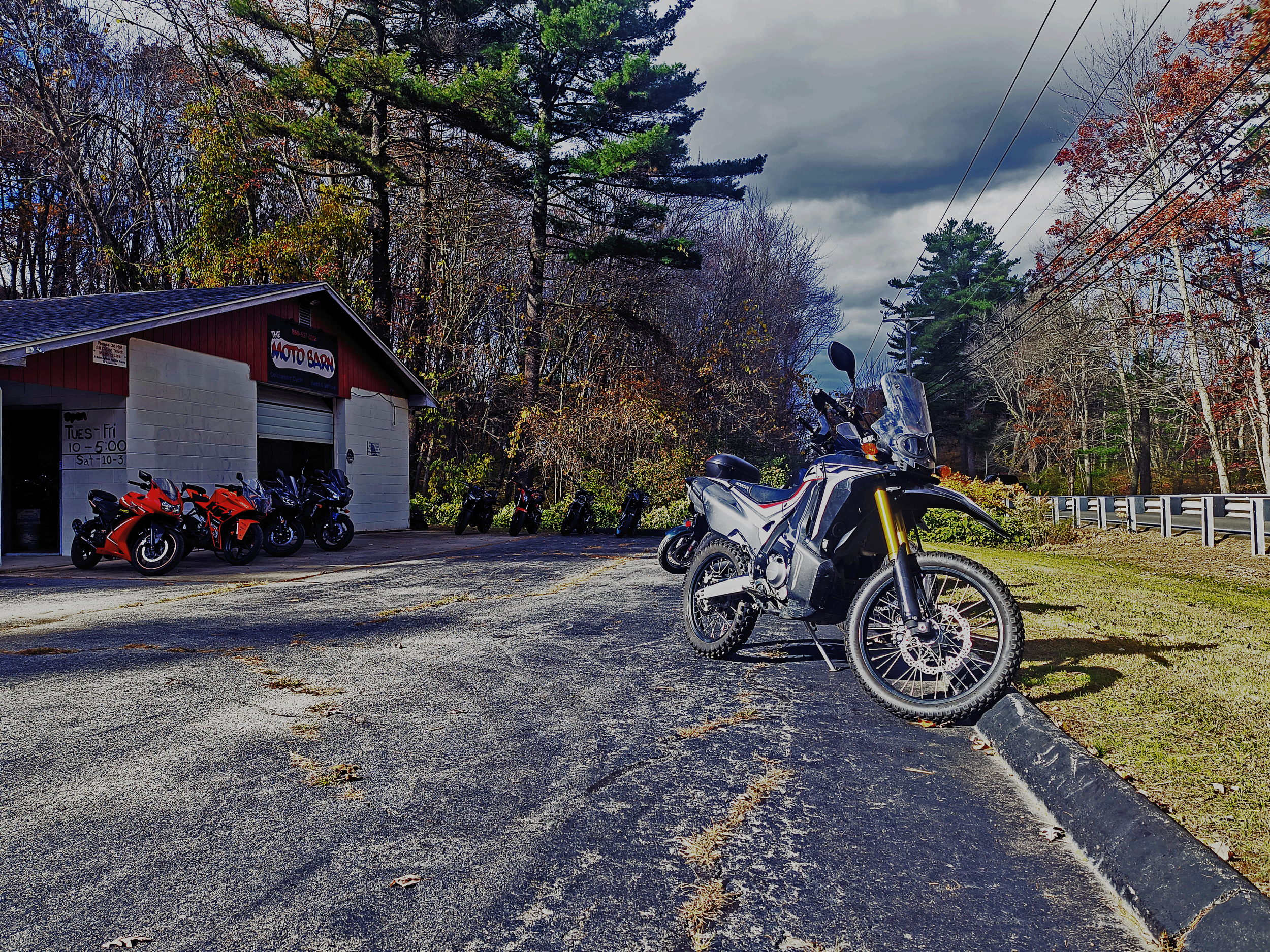 A motorcycle dealership with multiple motorcycles parked outside and a large adventure motorcycle in the foreground on a raised parking space, surrounded by trees and power lines under cloudy skies.