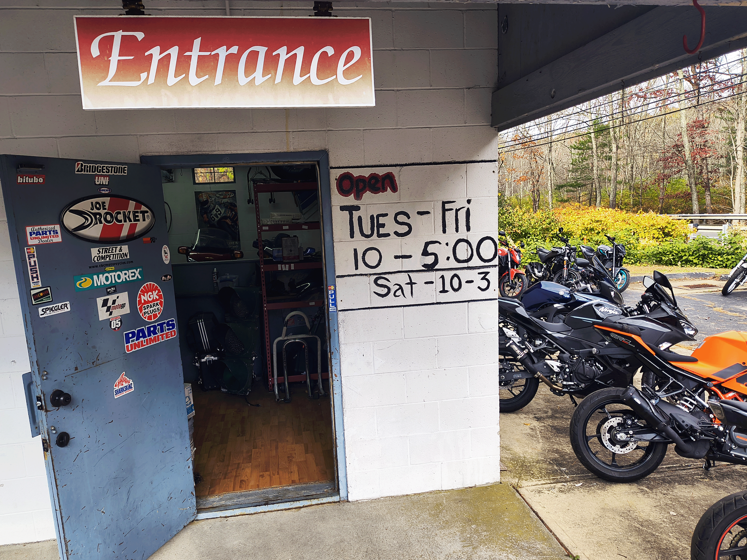 Outside motorcycle shop with an open door and a white wall displaying operating hours: Tuesday to Friday 10 AM to 5 PM, Saturday 10 AM to 3 PM; a parking lot filled with black and orange motorcycles; trees with autumn foliage in the background.