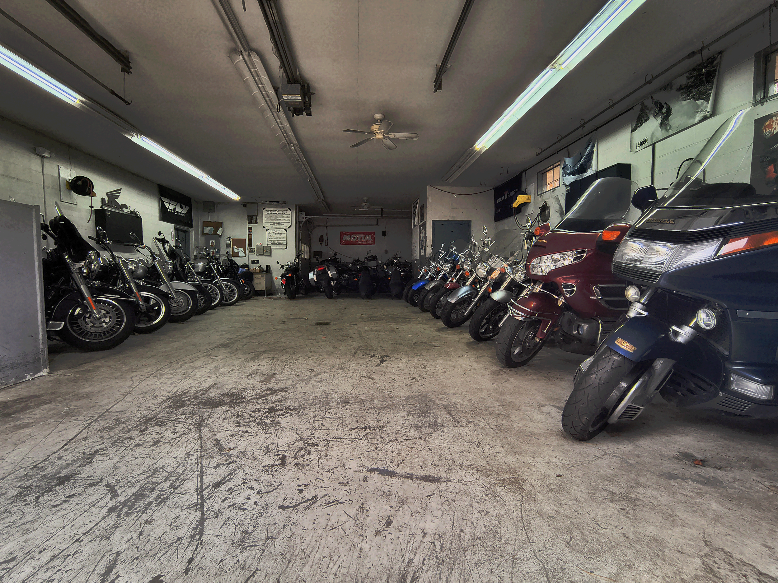 Indoor motorcycle garage with multiple parked bikes, including cruisers, touring bikes, and a trike, with tools and equipment on the walls.