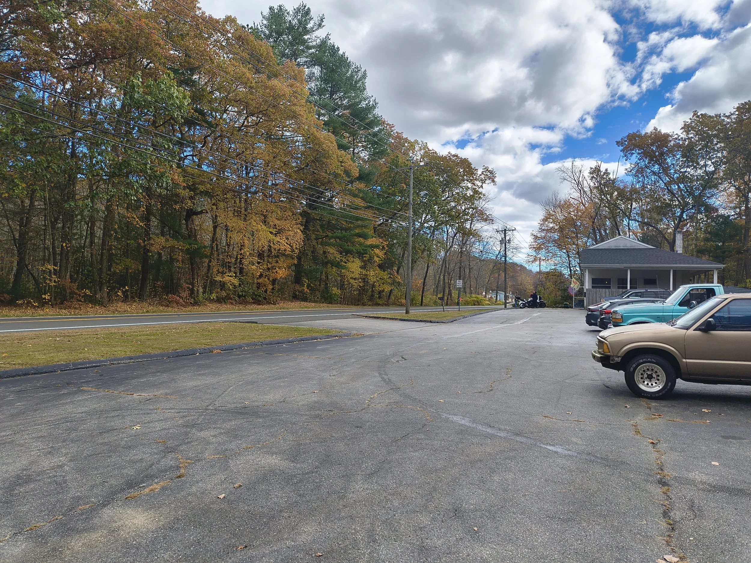 Parking lot with several parked cars and a small building, trees with autumn foliage on the left, and cloudy blue sky.