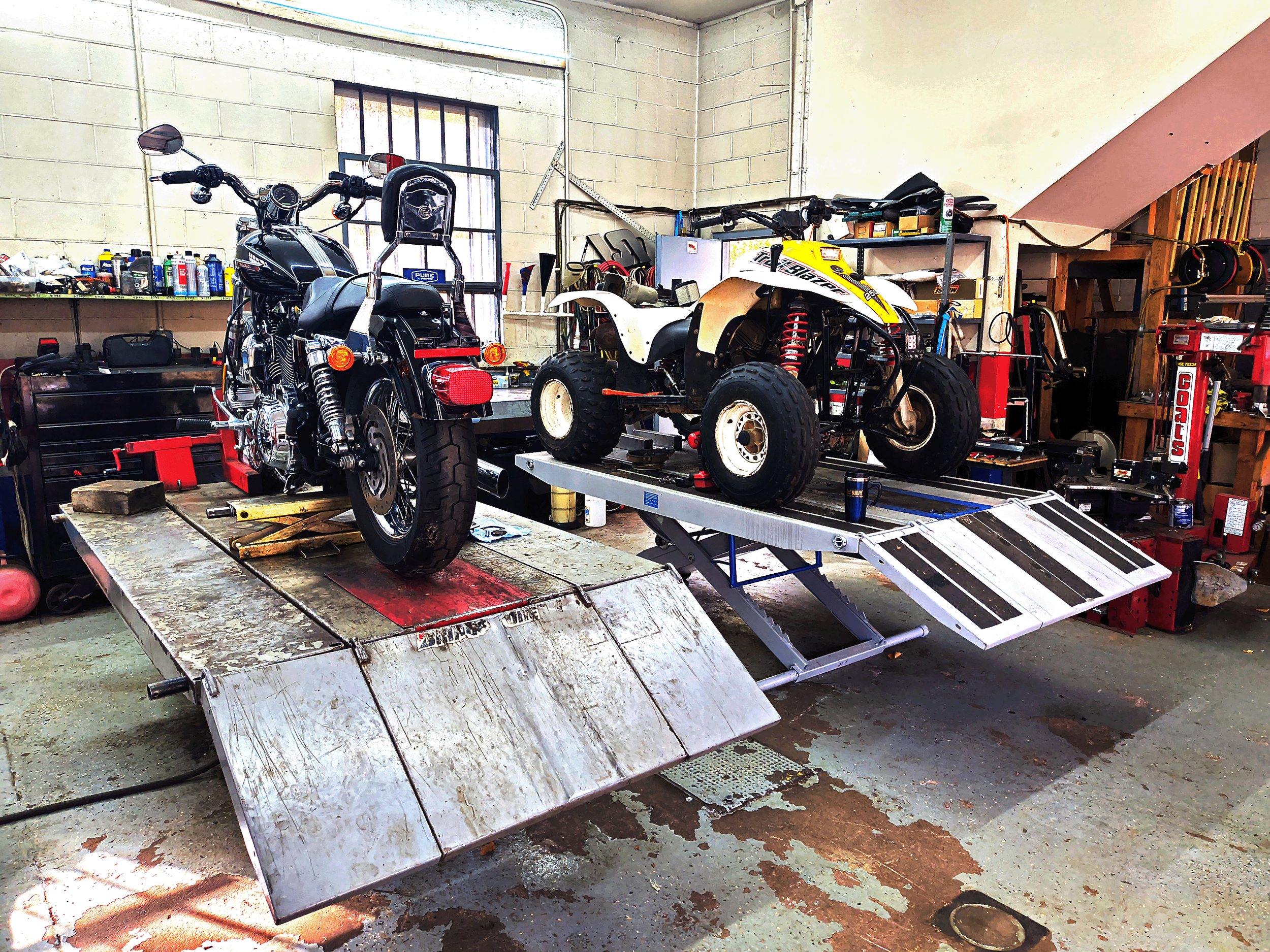 A motorcycle and a quad bike are on a vehicle lift in a workshop. The motorcycle is black with chrome accents, and the quad bike is yellow and white with large tires. The workshop has tools, bottles, and equipment on shelves and workbenches.