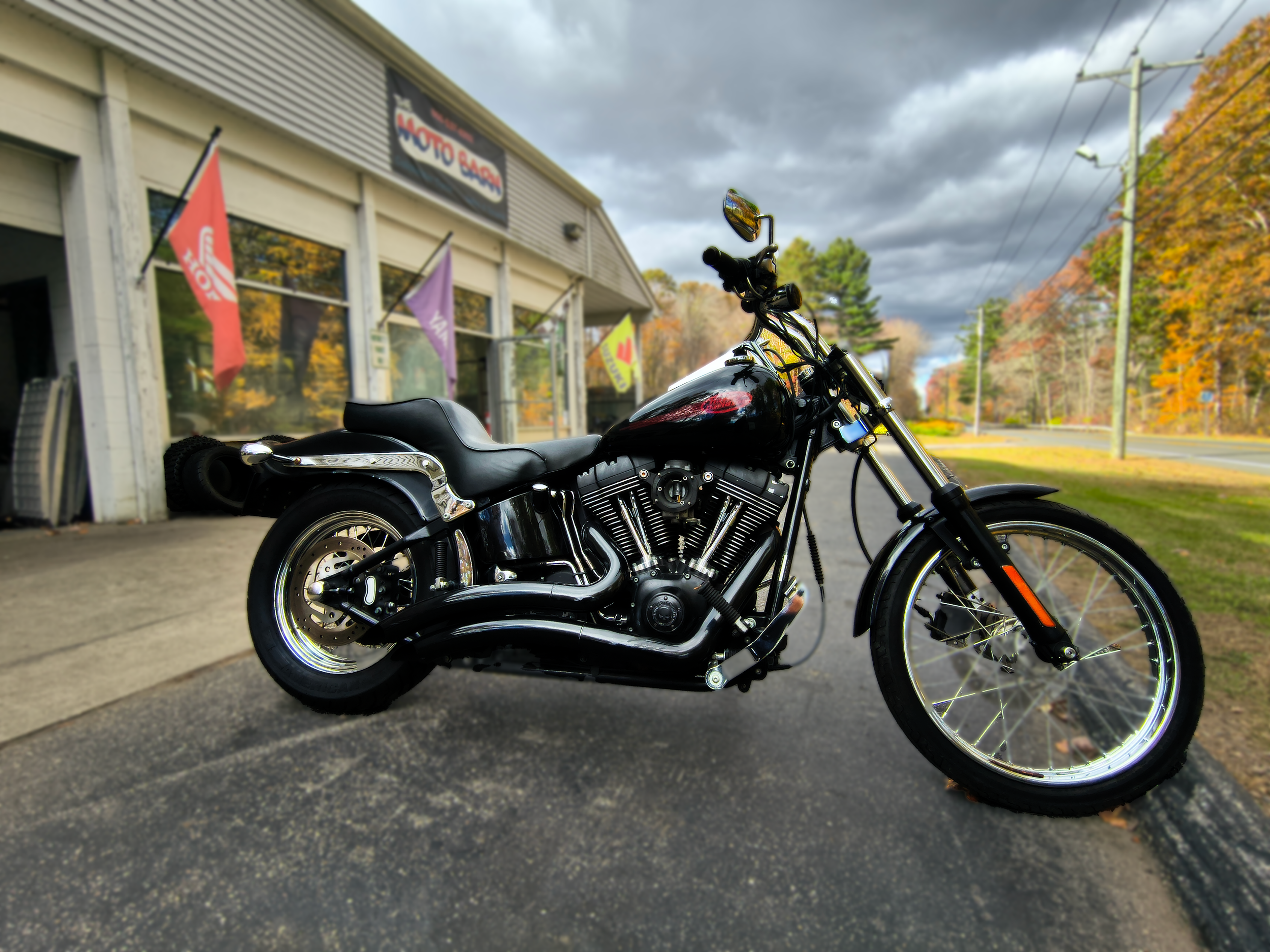 Black cruiser motorcycle parked outside motorcycle shop with flags and trees displaying fall foliage in the background.