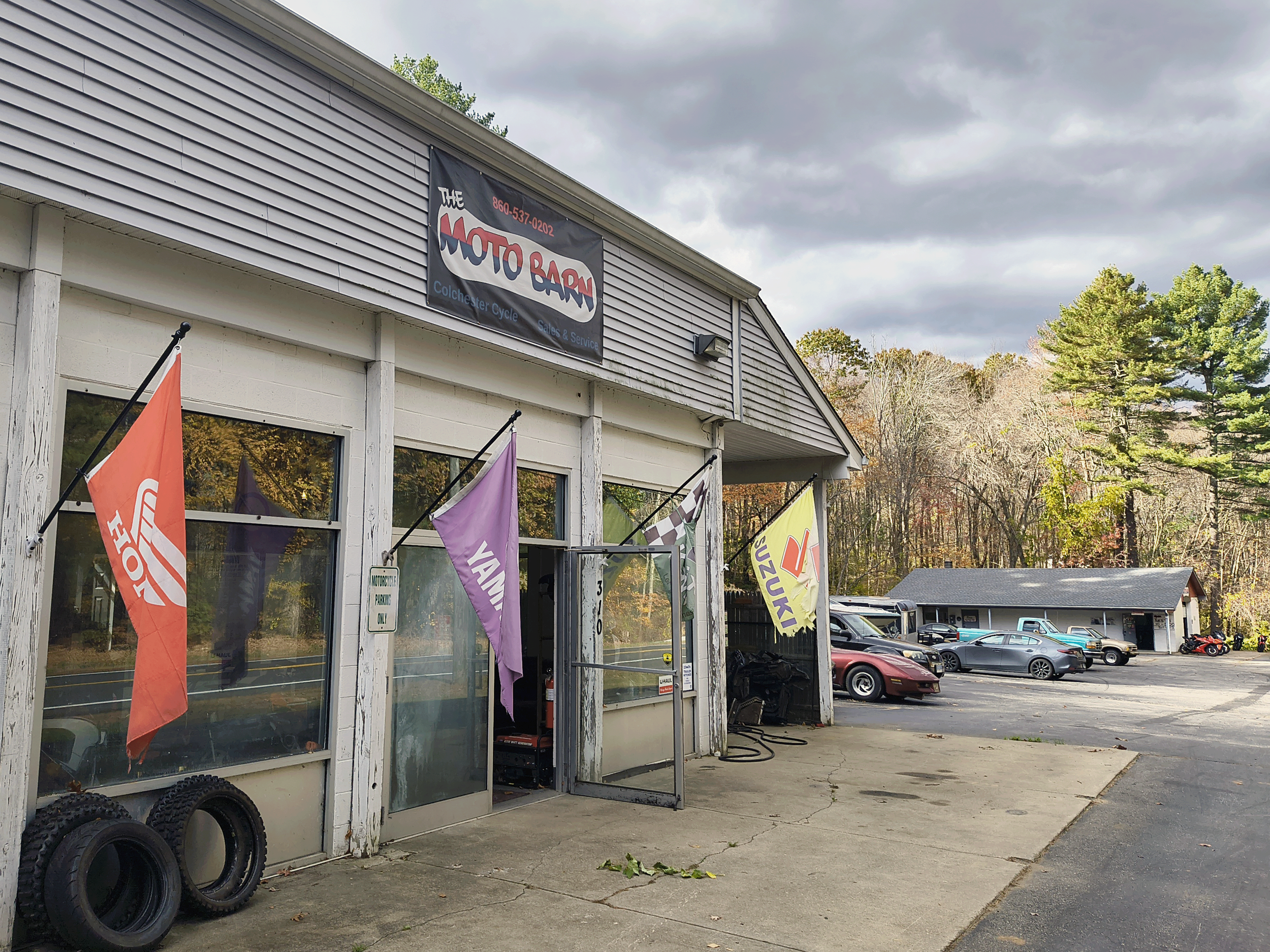 Exterior of a motorcycle shop named The Moto Barn, with flags displaying brands like Honda, Yamaha, and Suzuki, parked cars, and a wooded area in the background under cloudy skies.