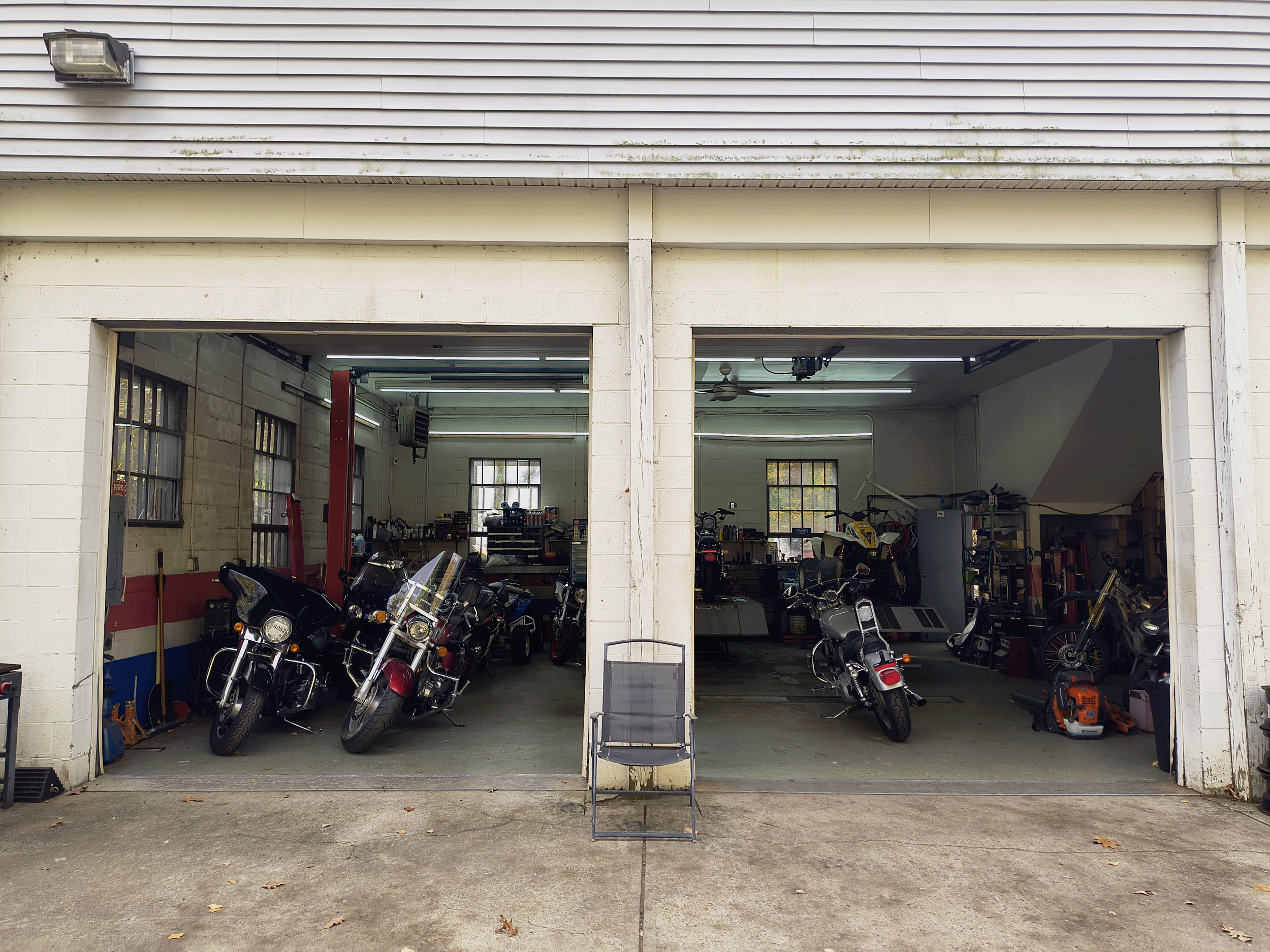 Inside a garage with motorcycles and motorcycle tools, with open garage doors, and a shopping chair placed outside.