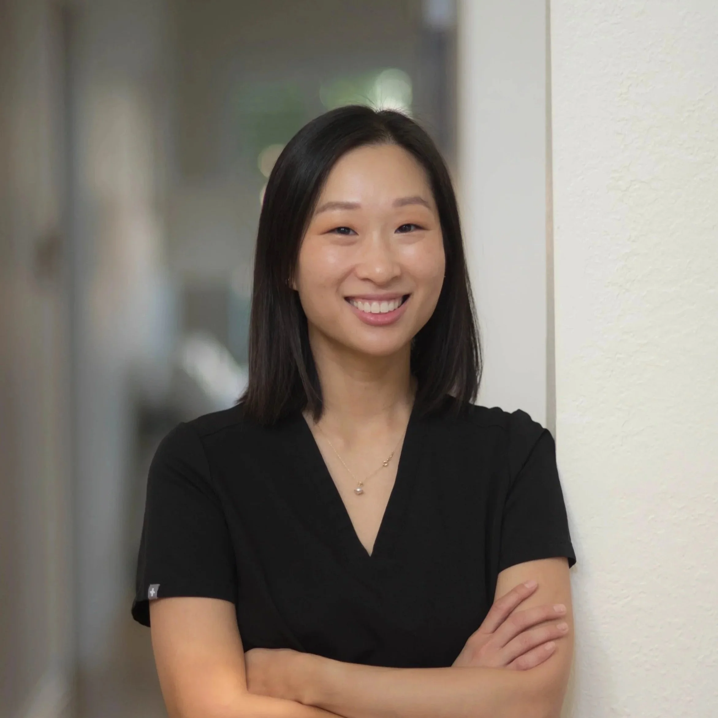 Smiling woman with shoulder-length black hair, wearing black scrubs and necklace, standing.