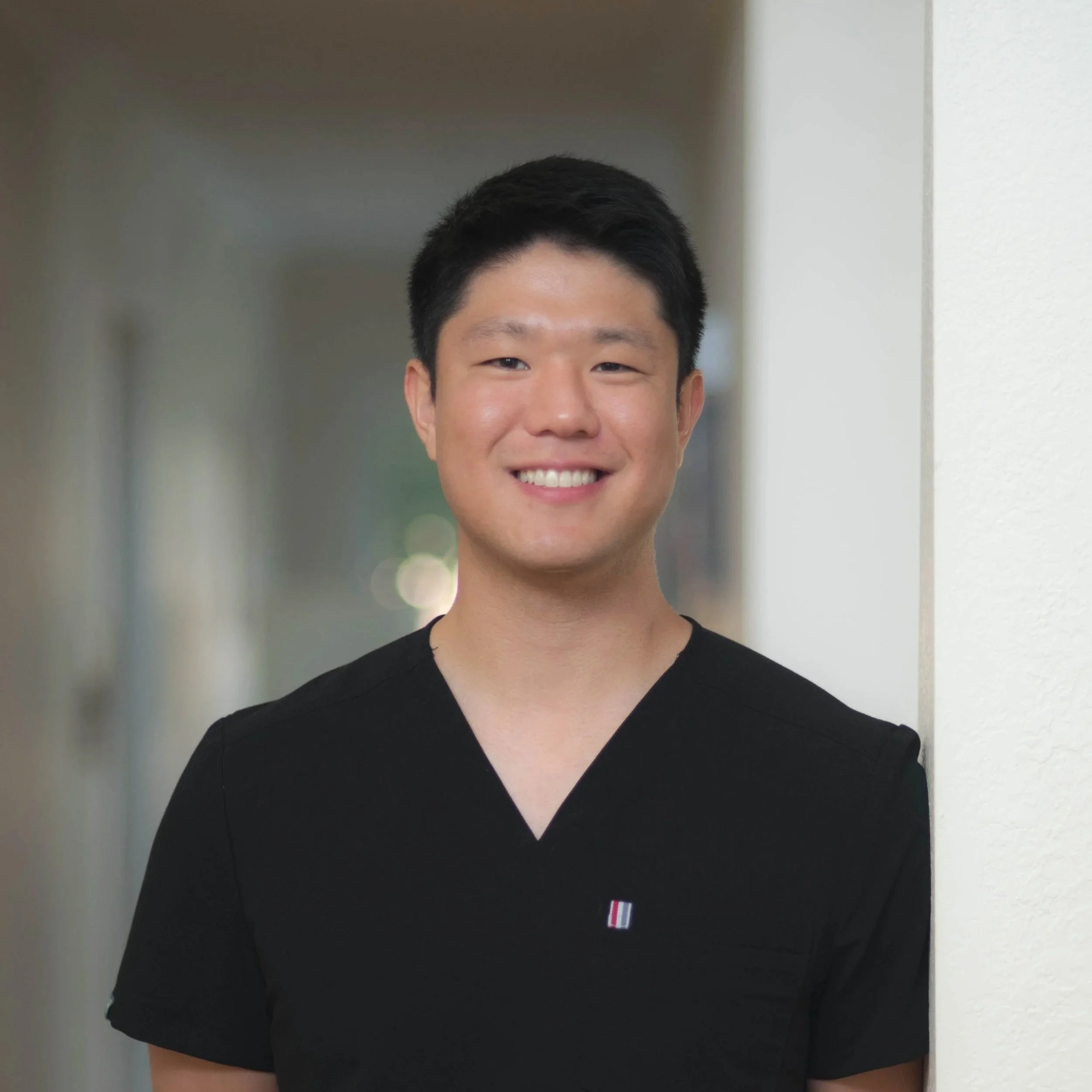 A smiling young man with short black hair, wearing a black V-neck top with a small logo, standing against a light-colored wall in an indoor hallway.