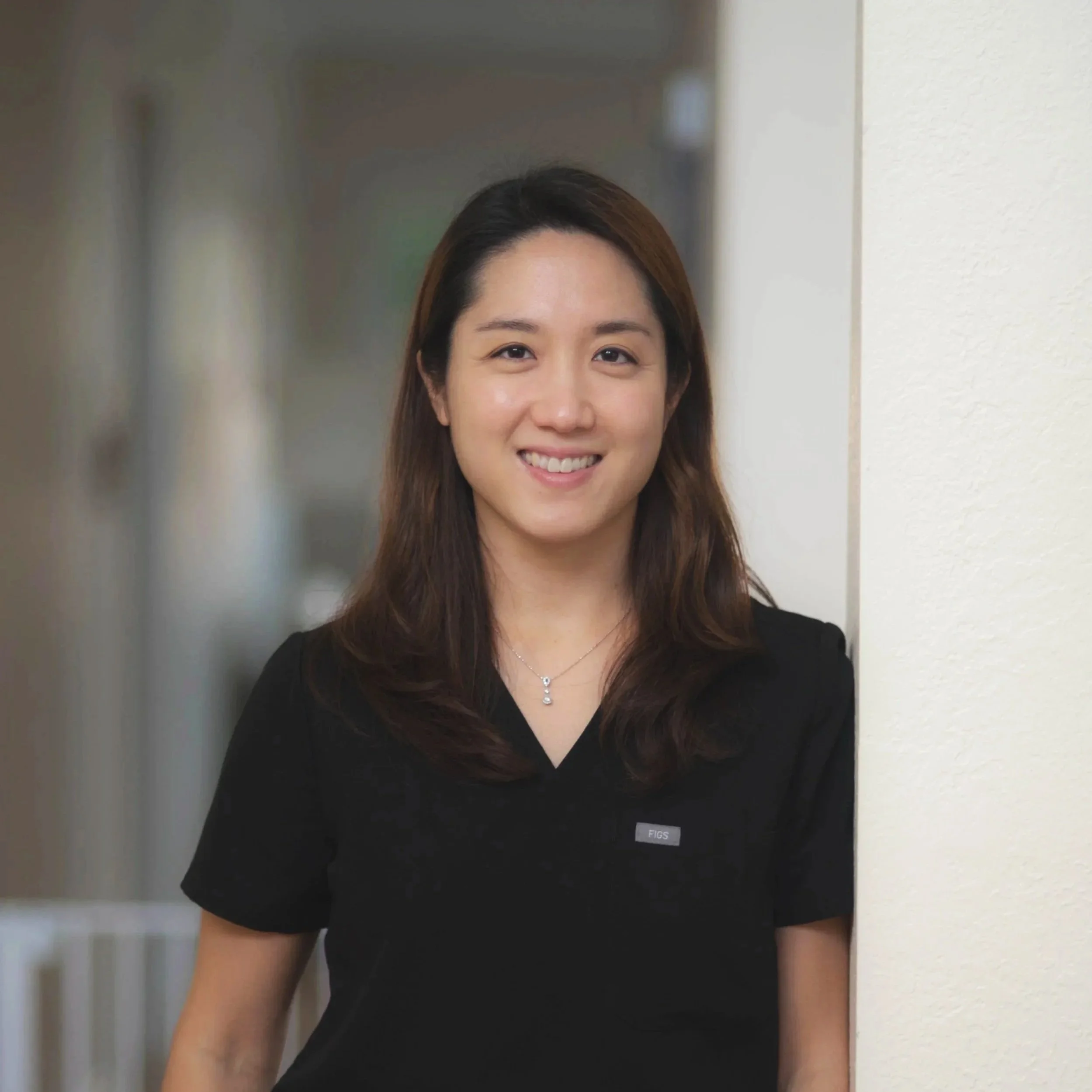 A smiling woman with shoulder-length brown hair, wearing a black top and a necklace, standing indoors next to a white wall.