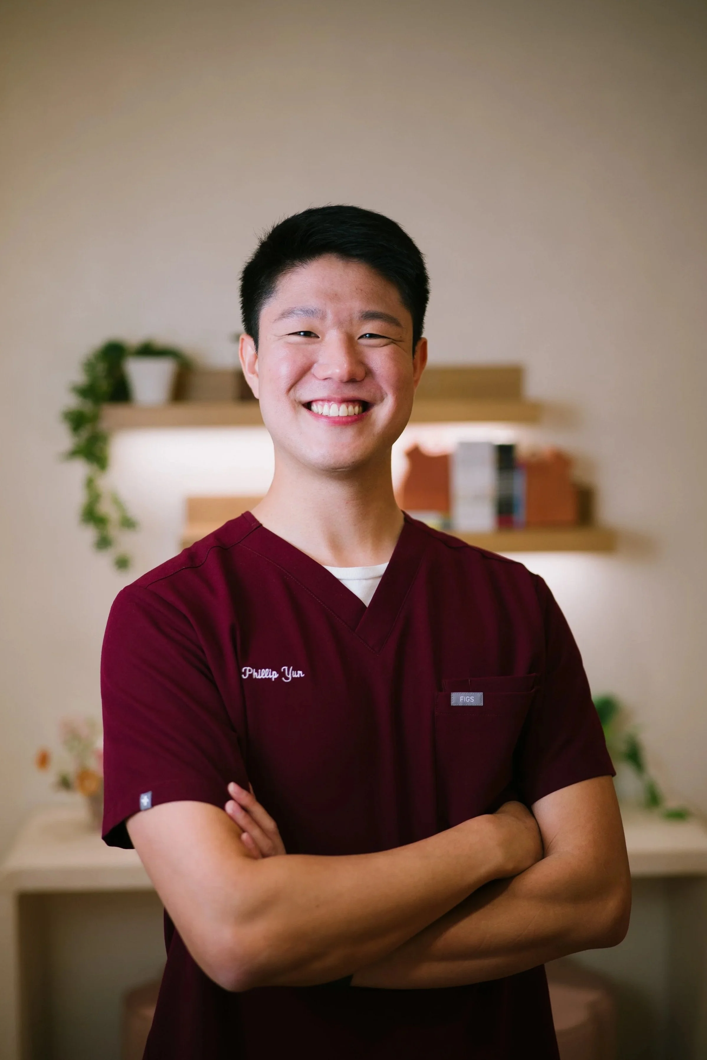 A smiling young man with short black hair, wearing a black V-neck top with a small logo, standing against a light-colored wall in an indoor hallway.