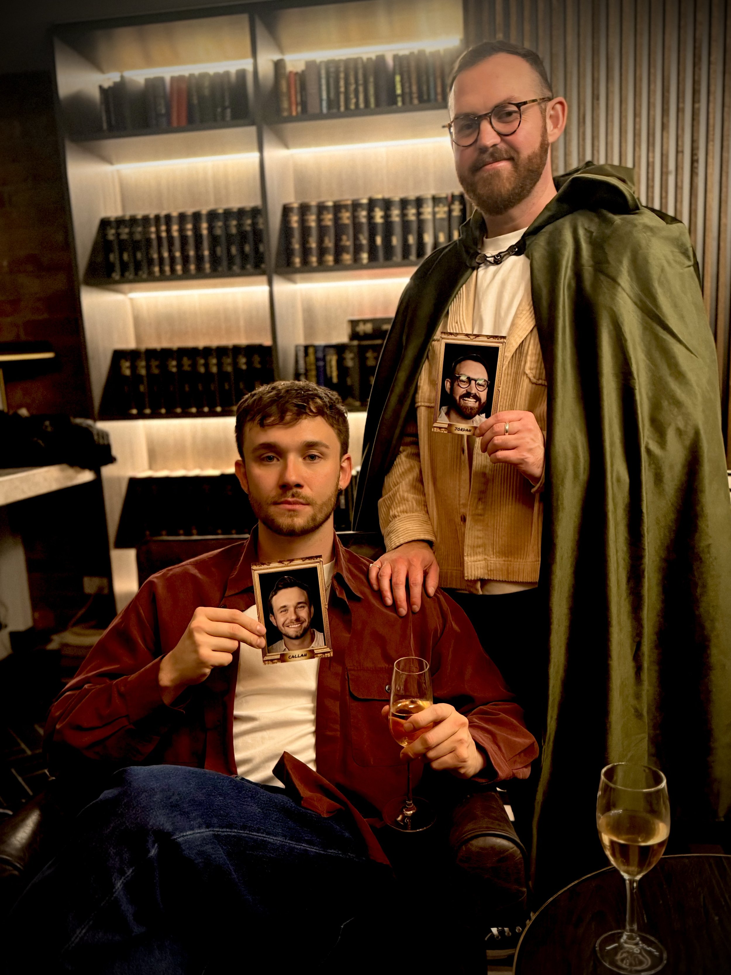 Two men posing in a cozy room with a bookshelf in the background. The man sitting on a chair is holding a picture of himself and a glass of wine, while the man standing behind him is holding a picture of the seated man. Both men have beards and are smiling slightly.