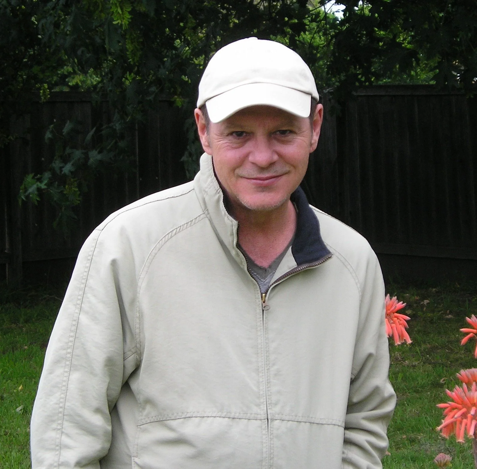 A man is standing outdoors in a garden with green grass and flowers, wearing a beige cap and a beige jacket, smiling slightly. Tony Stoufer, Nashville Bound, Lonely Just Goes On, Broken Heart Surgery, Stormy Night, Little Darli', Maybe Not, COVID 19