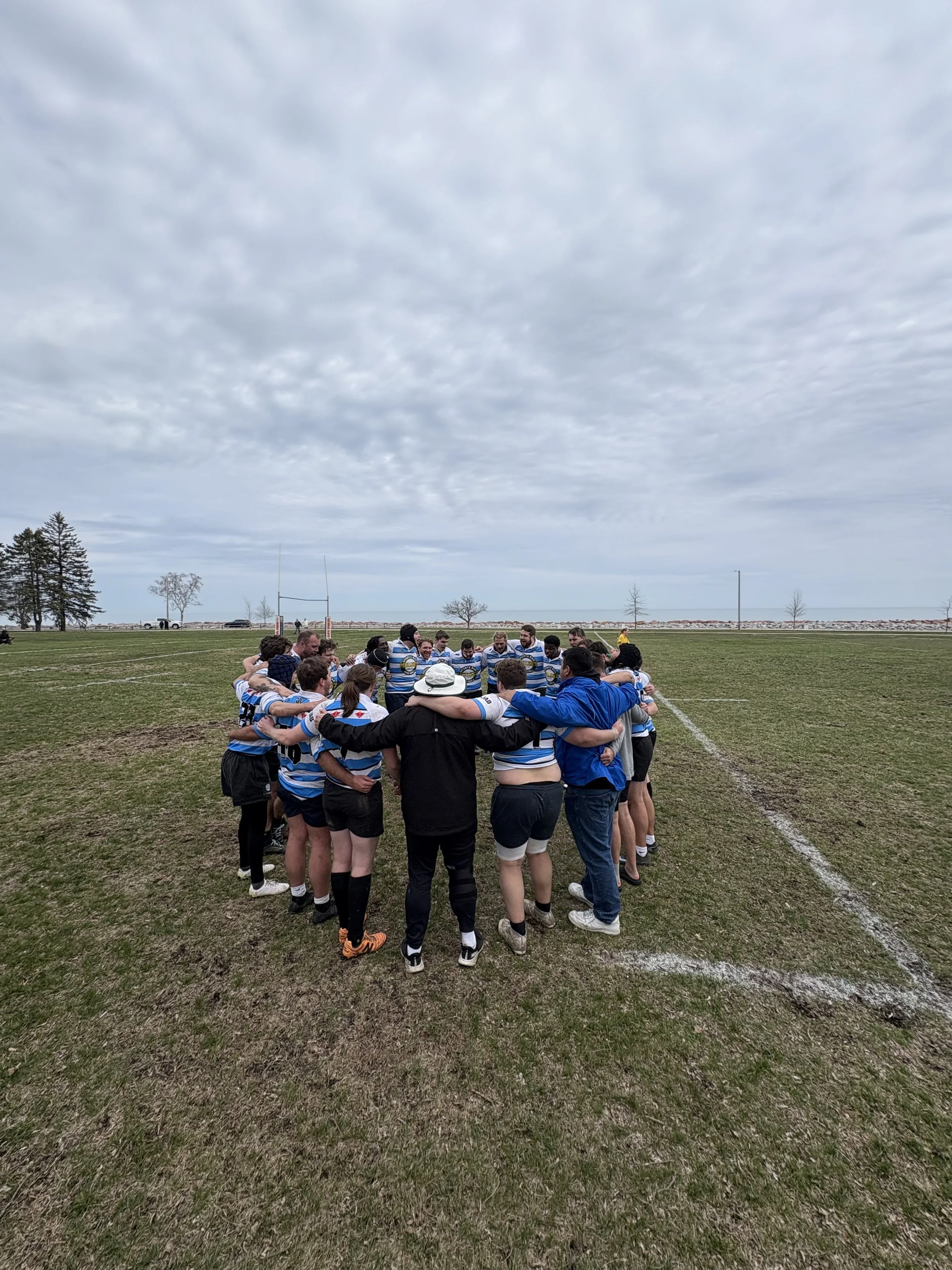 A group of people standing in a circle on a grassy field, huddled with arms around each other's shoulders during daytime with cloudy sky.