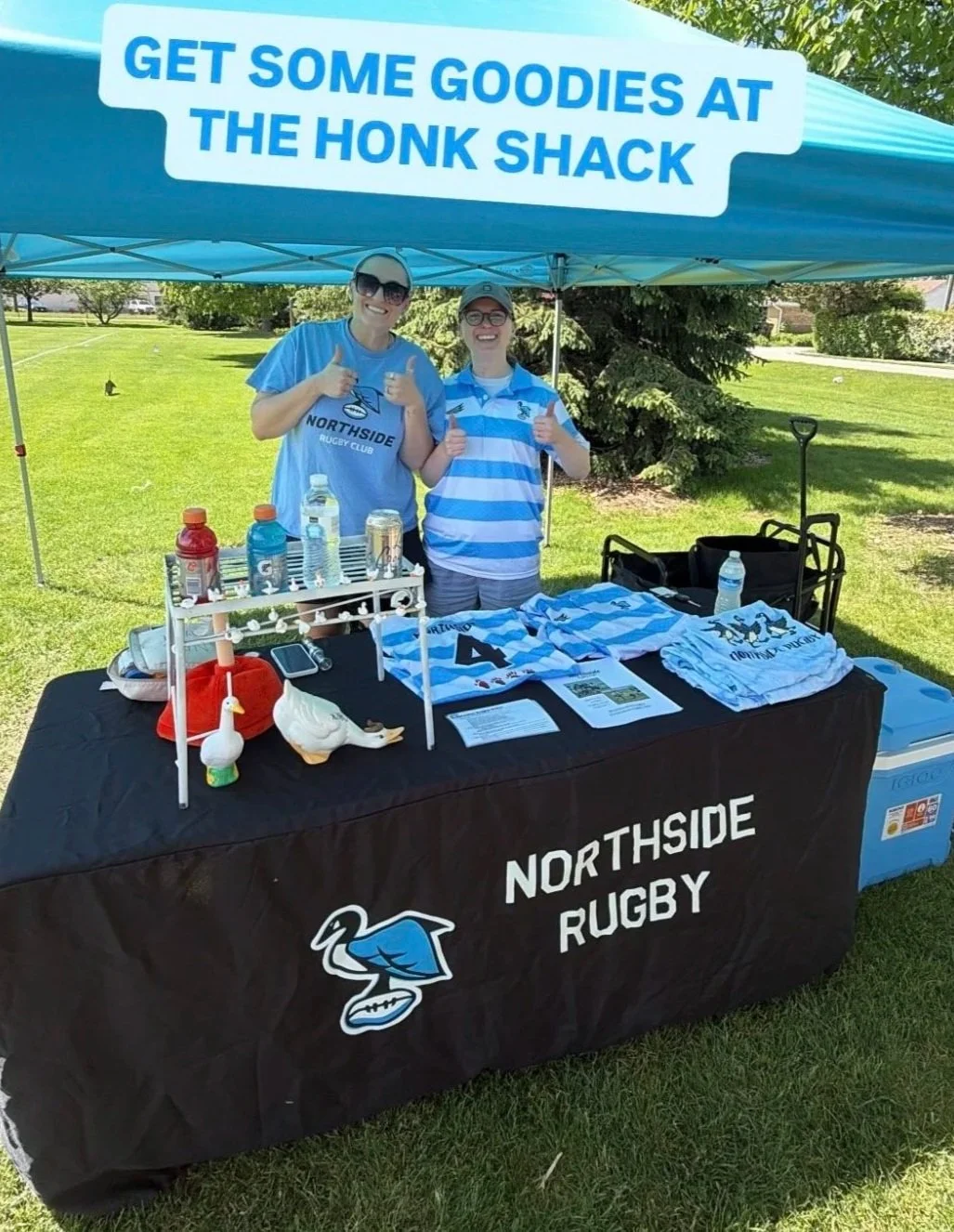 Two women standing behind a table under a blue canopy with a sign that says, 'Get Some Goodies at The Honk Shack.' They are smiling and giving thumbs up. The table is covered with Northside Rugby merchandise, including t-shirts, and athletic gear. The scene is outdoors on a sunny day in a park.