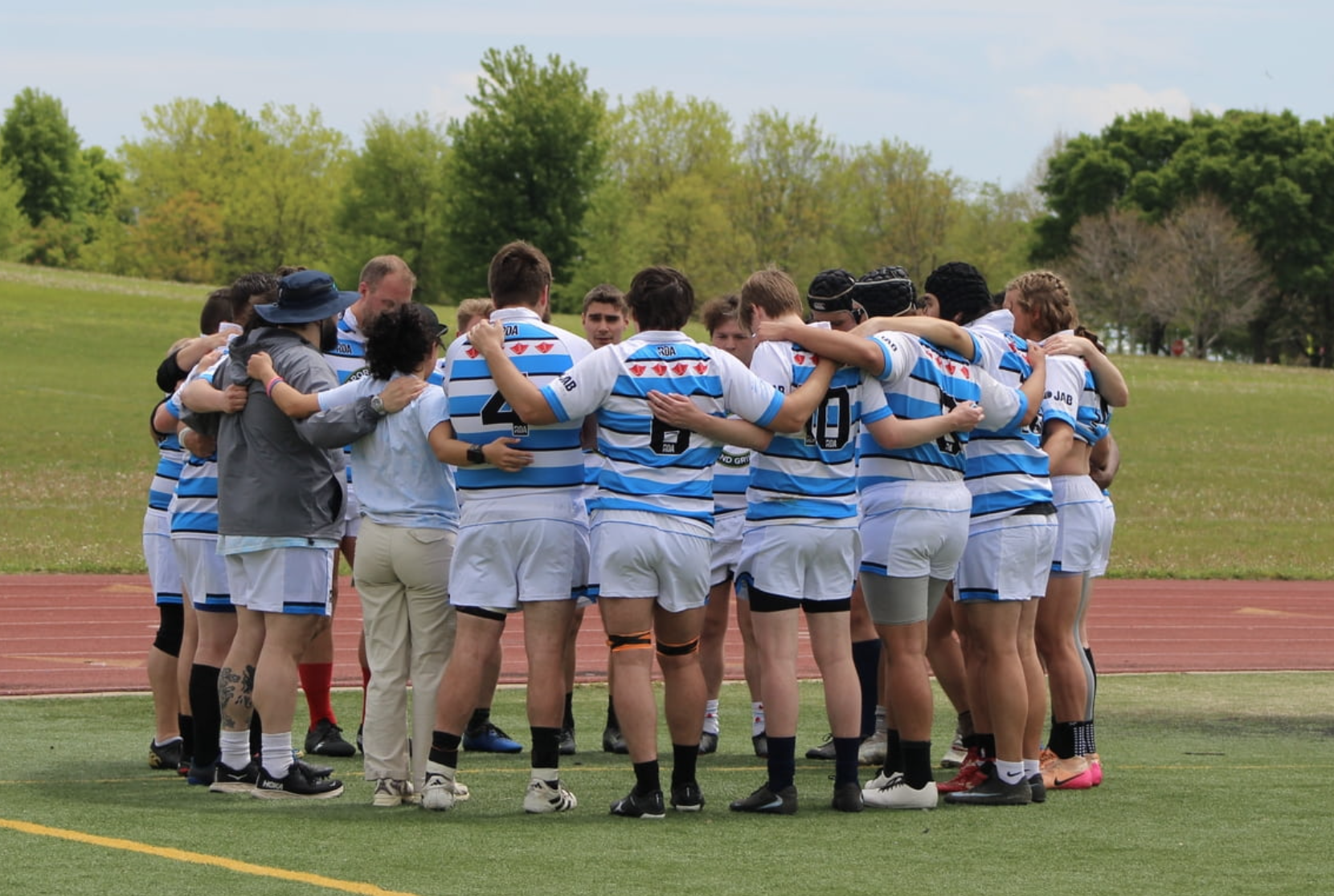 A group of women and men rugby players in white and blue jerseys huddle together on a field, embracing each other, during a game or practice, with a green park and trees in the background.