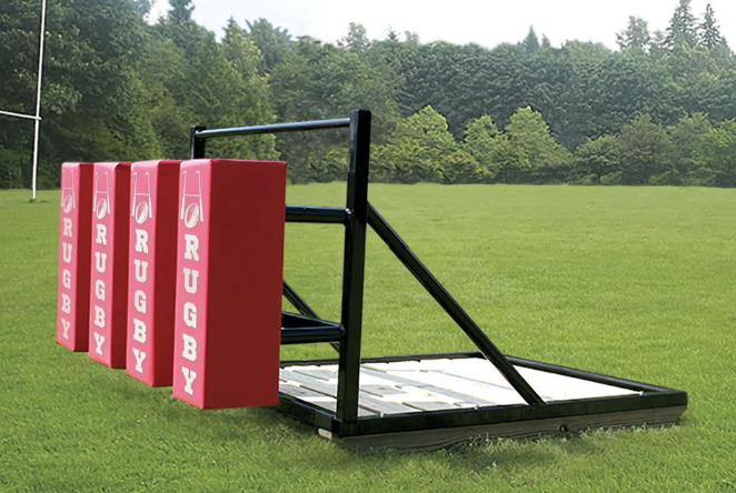 A rugby-themed cornhole game set up on a grassy field with a backdrop of trees. The game features pink boards with the word 'RUGBY' written vertically in white.