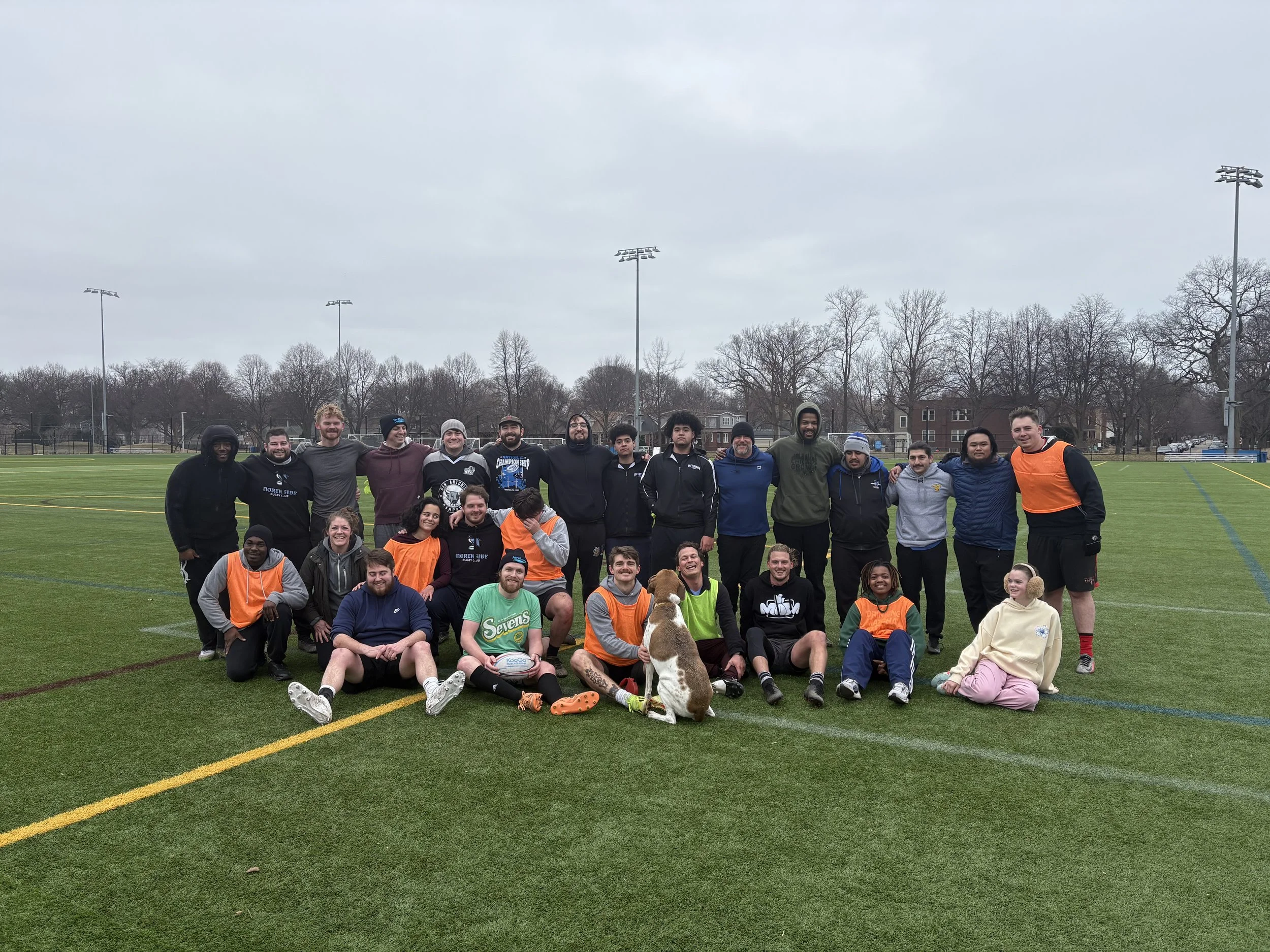 A group of people and a dog on a sports field during daytime. The group includes men, women, and children, some sitting and some standing. The background shows leafless trees, a few buildings, and overcast skies.