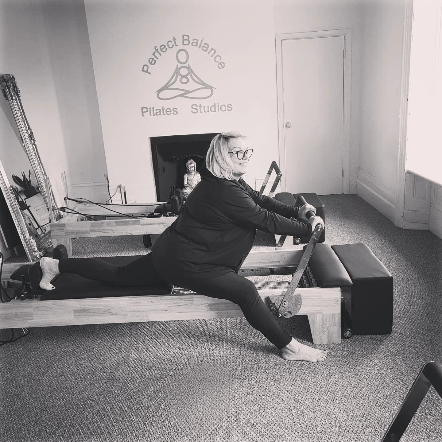 A woman with glasses and shoulder-length hair is doing a Pilates exercise on a reformer machine in a Pilates studio. The studio has a sign on the wall that says 'Perfect Balance Pilates Studios' and features a yoga pose logo. There is a mirror and a small statue of Buddha in the studio.