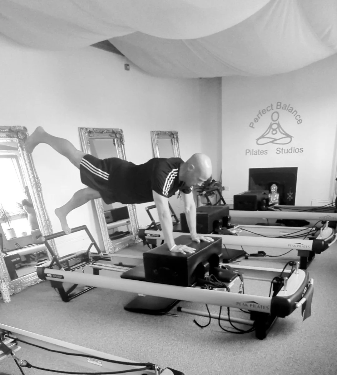 A man practicing Pilates on specialized reformer machines in a studio with mirrors and the sign 'Perfect Balance Pilates Studios' on the wall.