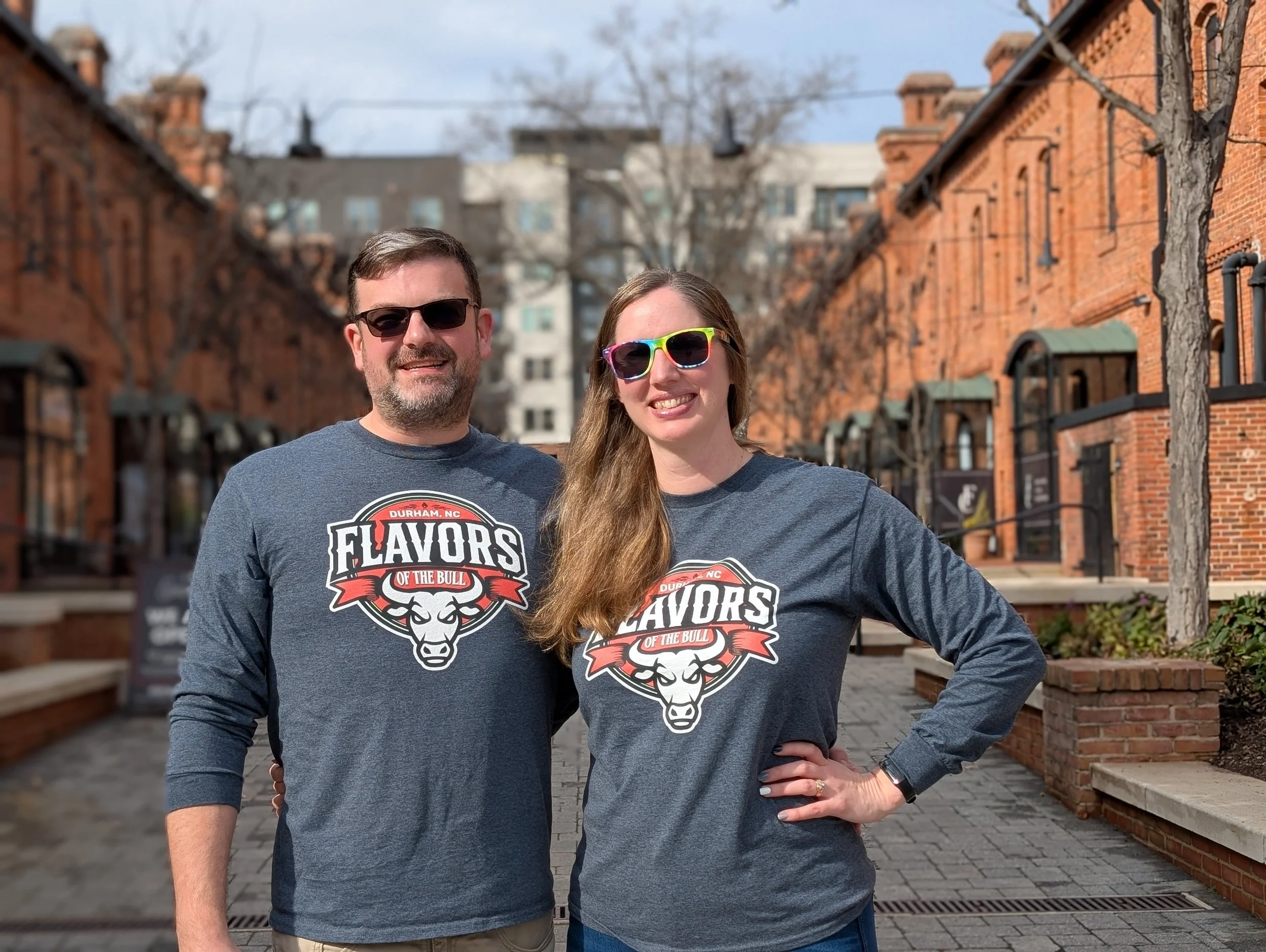 A man and woman standing outdoors on a brick-paved street lined with brick buildings, both wearing matching gray T-shirts with a bull logo and text that says 'Flavors of the Bull' and 'Durham, NC,' smiling at the camera, with clear weather and leafless trees in the background.