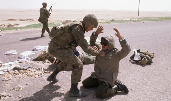 A soldier helps a civilian who is kneeling on the ground with his hands raised in a defensive gesture. Two other soldiers are in the background, walking along the road in a desert landscape.
